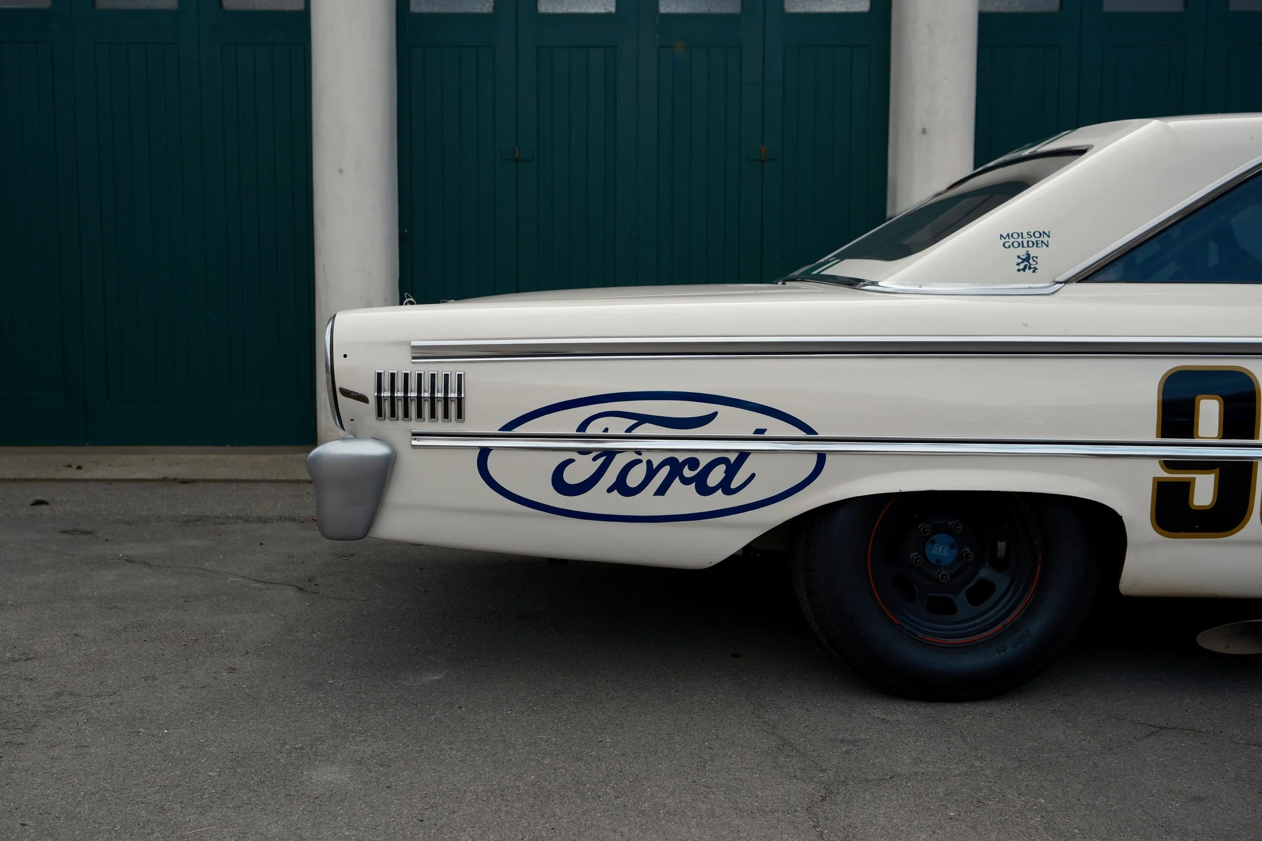 Close-up of a vintage white Ford race car with logos, parked on the street, in front of a green fence and concrete pillars.