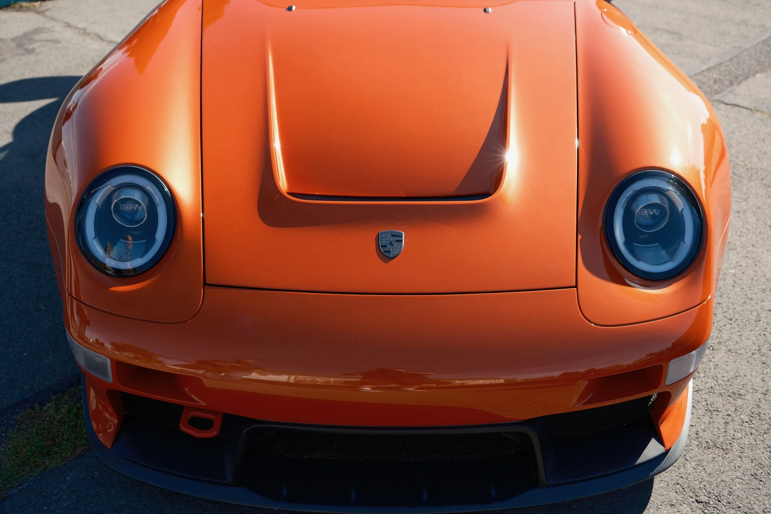 Front view of a bright orange Porsche sports car on a concrete surface with shadows from sunlight.