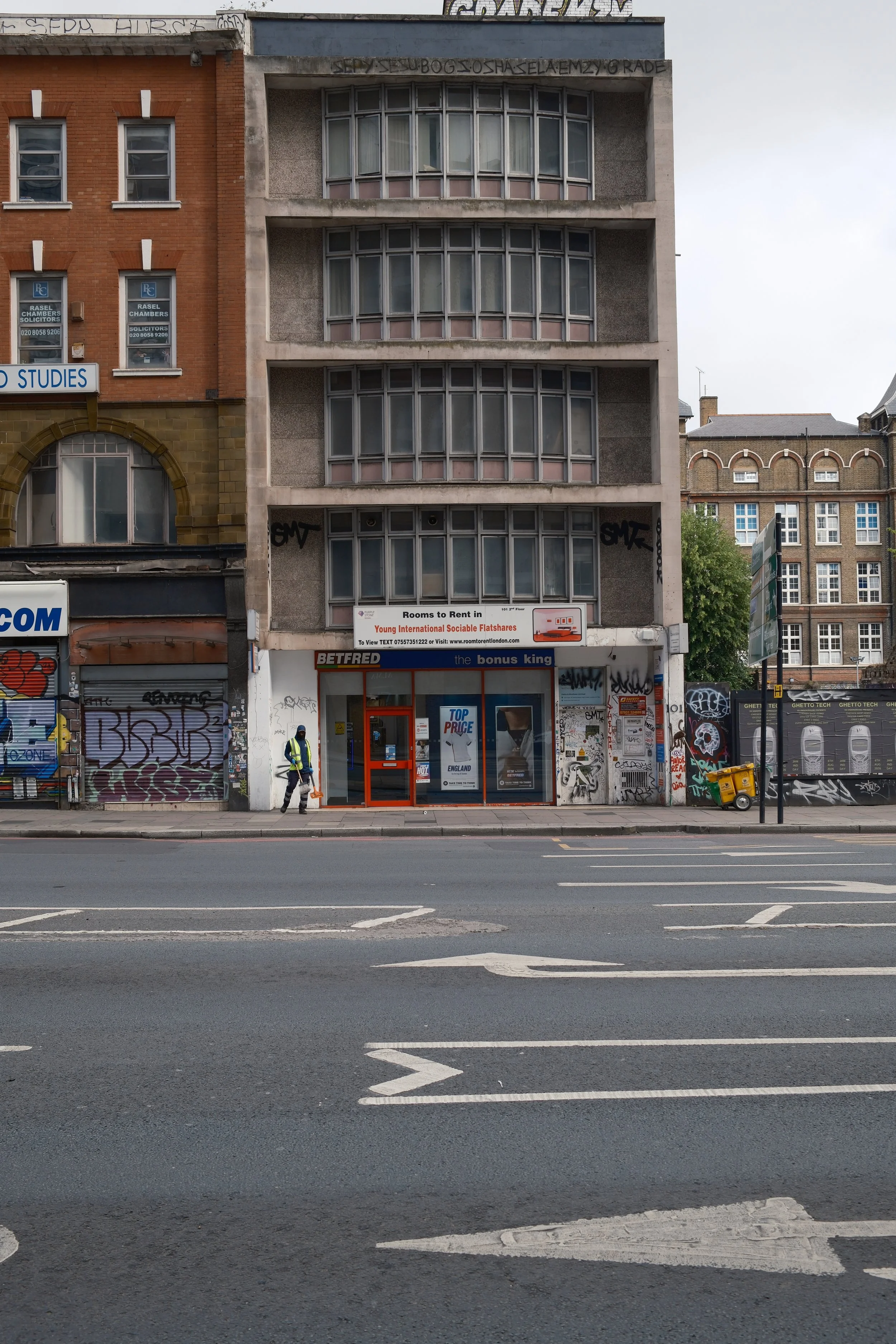 Urban street scene with a four-story building that has a retail store on the ground floor and residential or office spaces above. The store has a sign advertising rooms to rent, a betting shop inside, and is covered in graffiti. A person in a reflect