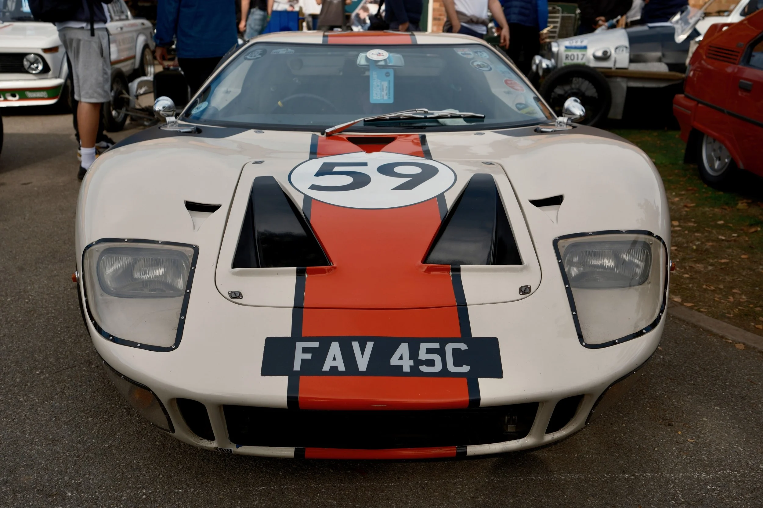 Front view of a vintage race car with the number 59 on the hood, parked at a car show with people and other classic cars in the background.