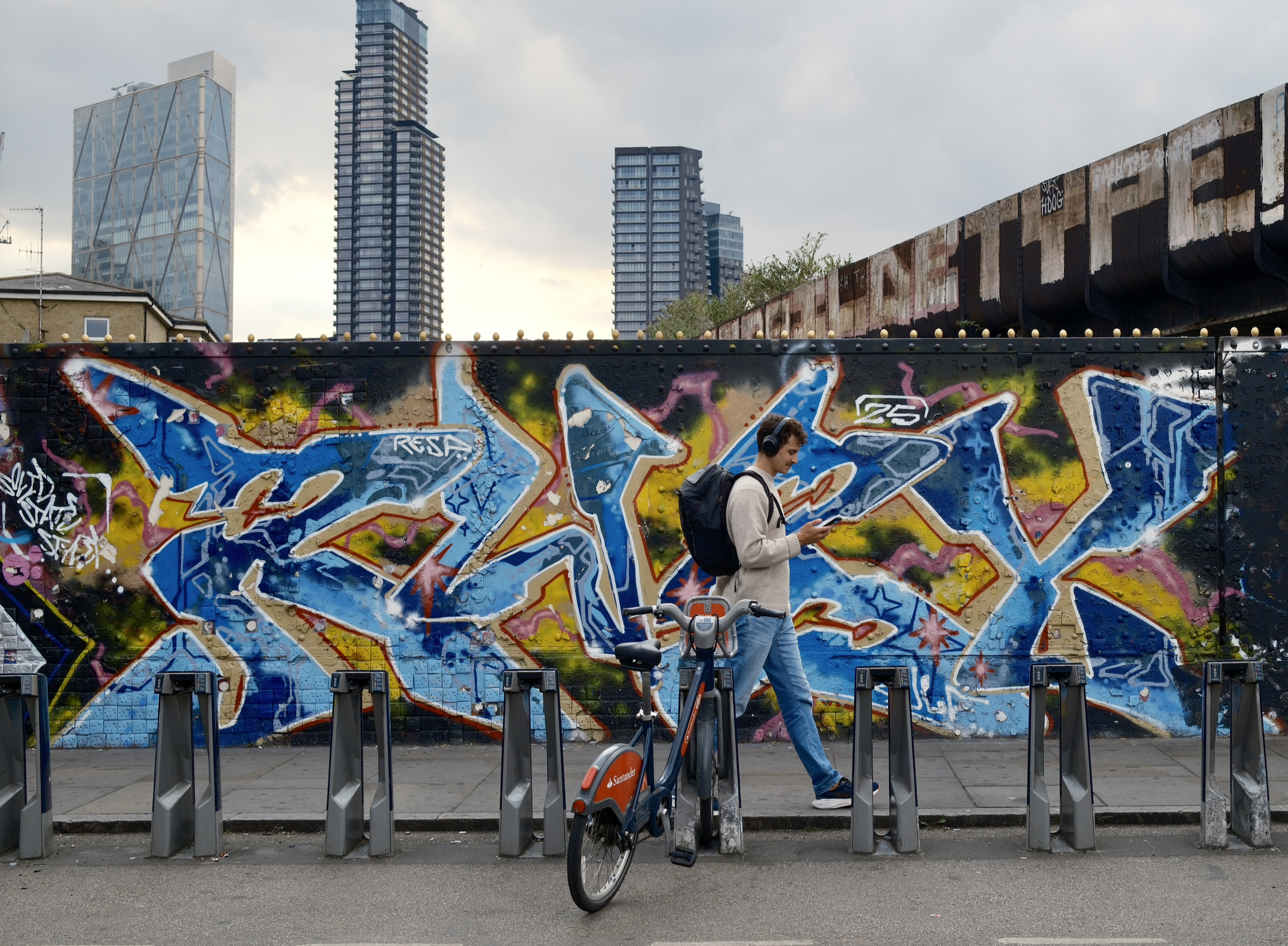 A young man with headphones, dressed in a gray hoodie and jeans, standing next to a bicycle and using a smartphone against a graffiti-covered wall with a cityscape of tall buildings in the background.