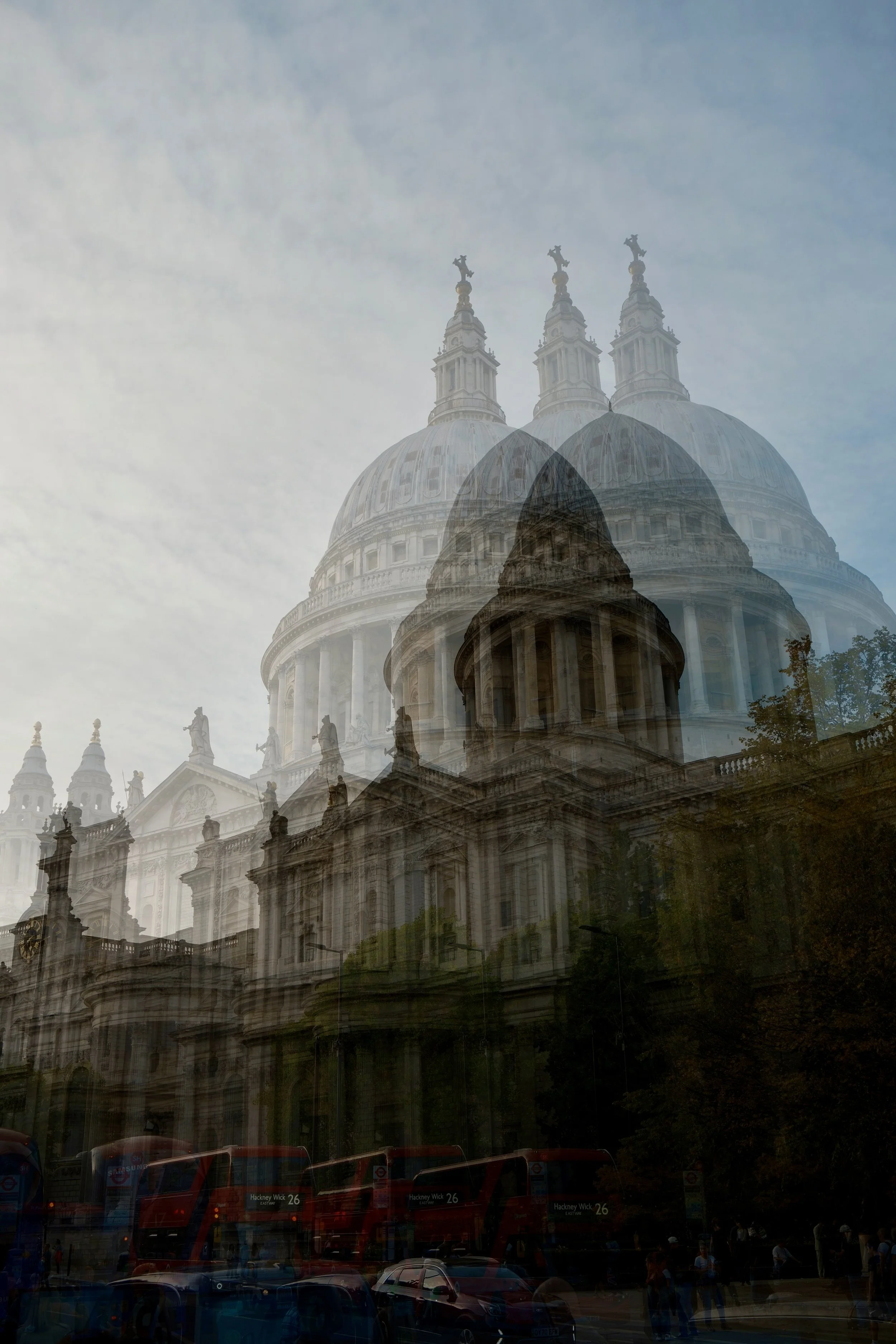 Multiple red double-decker buses and cars in front of a historic building with large domes and statues, under a cloudy sky.