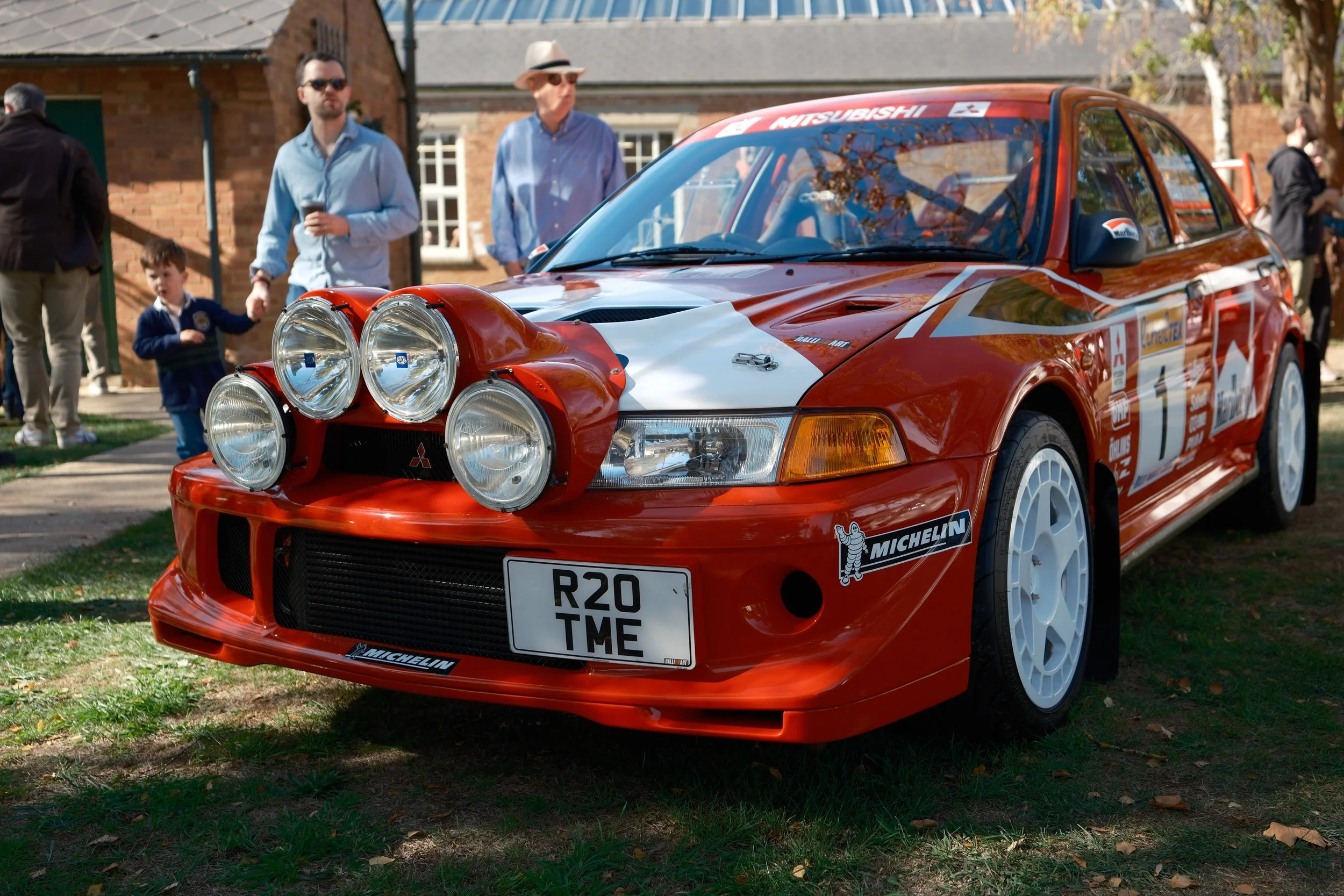 A red Mitsubishi rally car with multiple large front lights on the hood, parked on grass during an outdoor event. Several people, including children and adults, are standing nearby, some with drinks, in front of a brick building and trees.