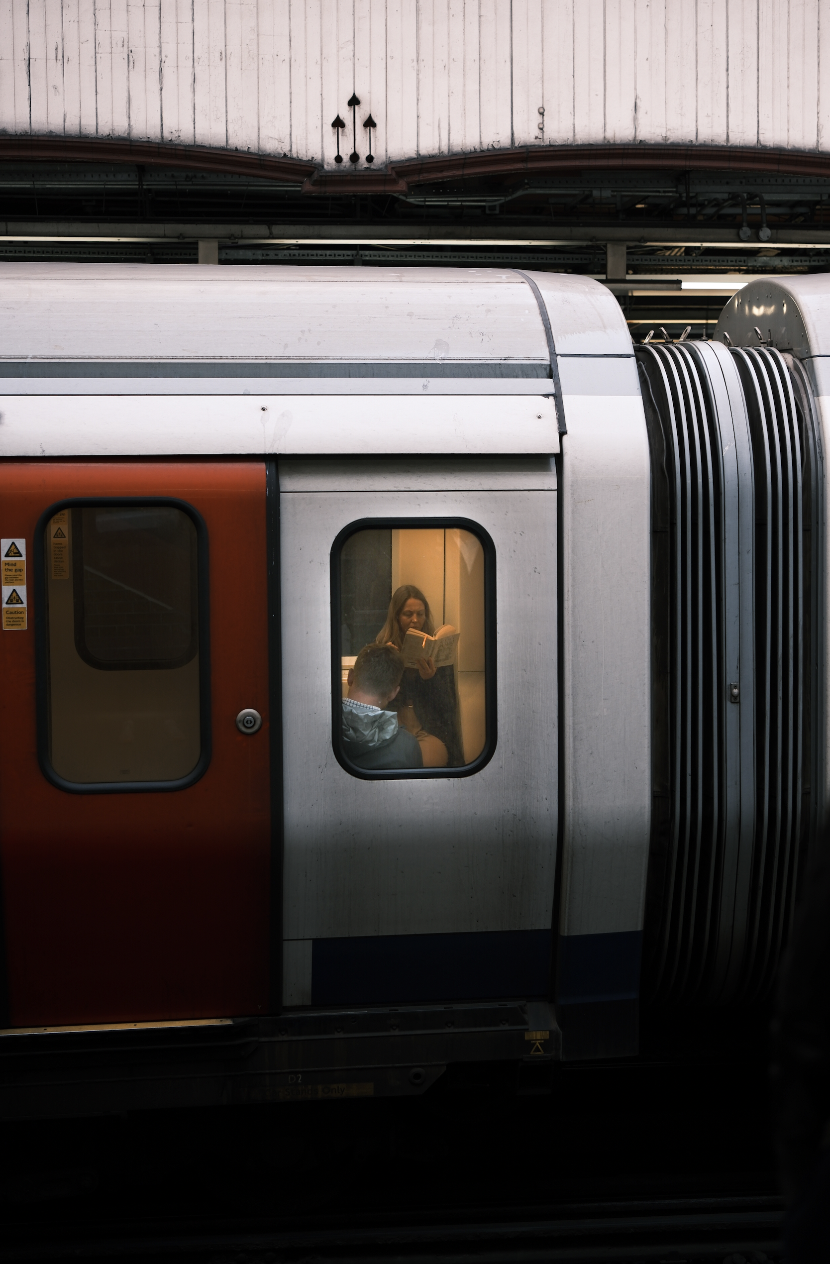 A train at a station with a woman reading a book and a person with a hooded jacket inside.