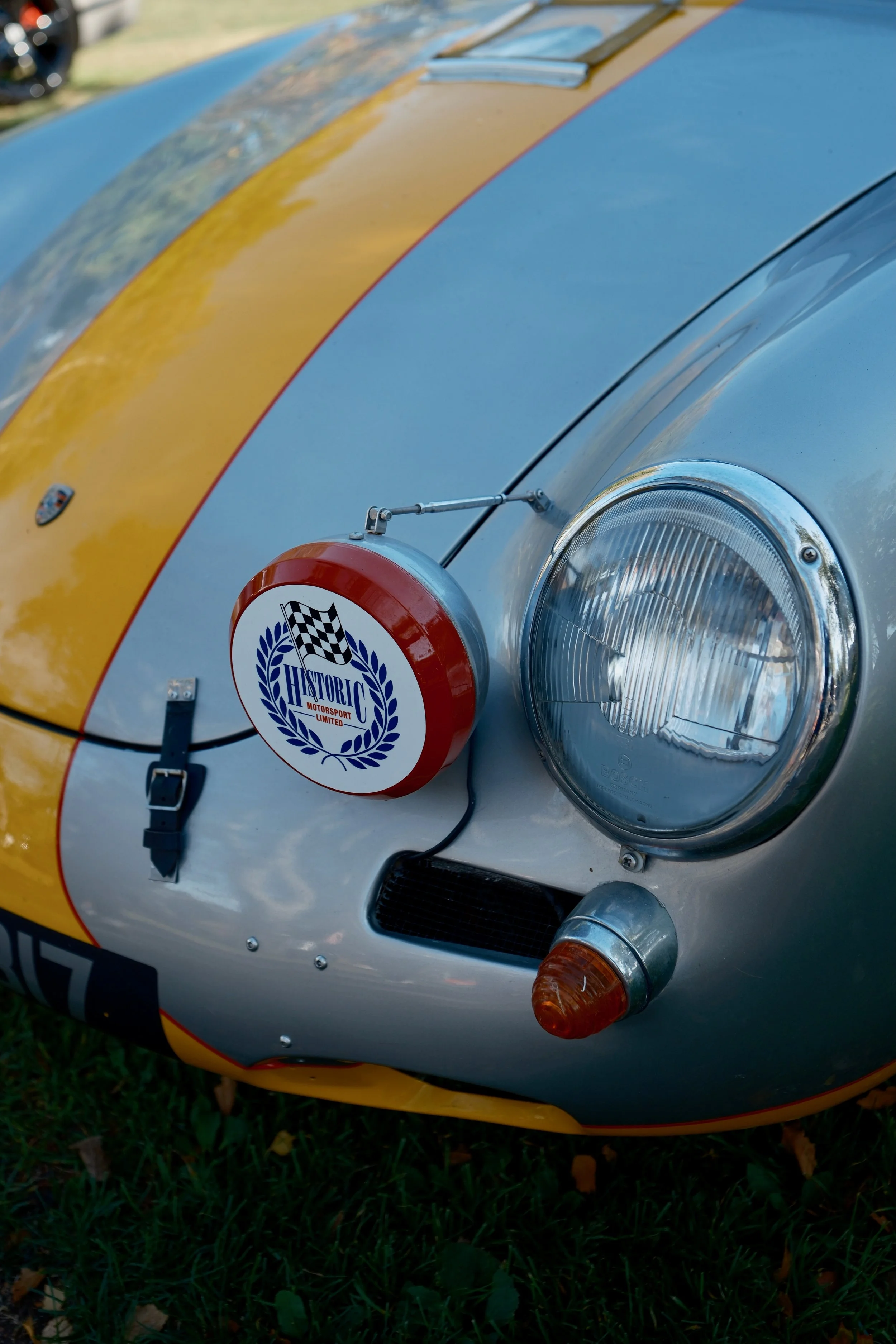 Close-up of the front of a silver racing car with yellow and red stripes, with a headlight, turn signal, and a badge that reads 'Historic Motorsport Limited.'