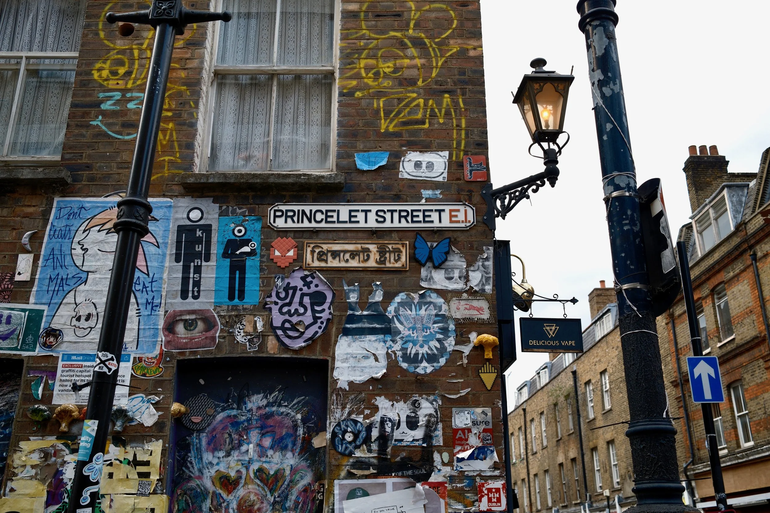 Wall covered with various street art, stickers, posters, and graffiti, with a street sign reading 'PRINCELET STREET E.I.' and a decorative black lamppost in front.
