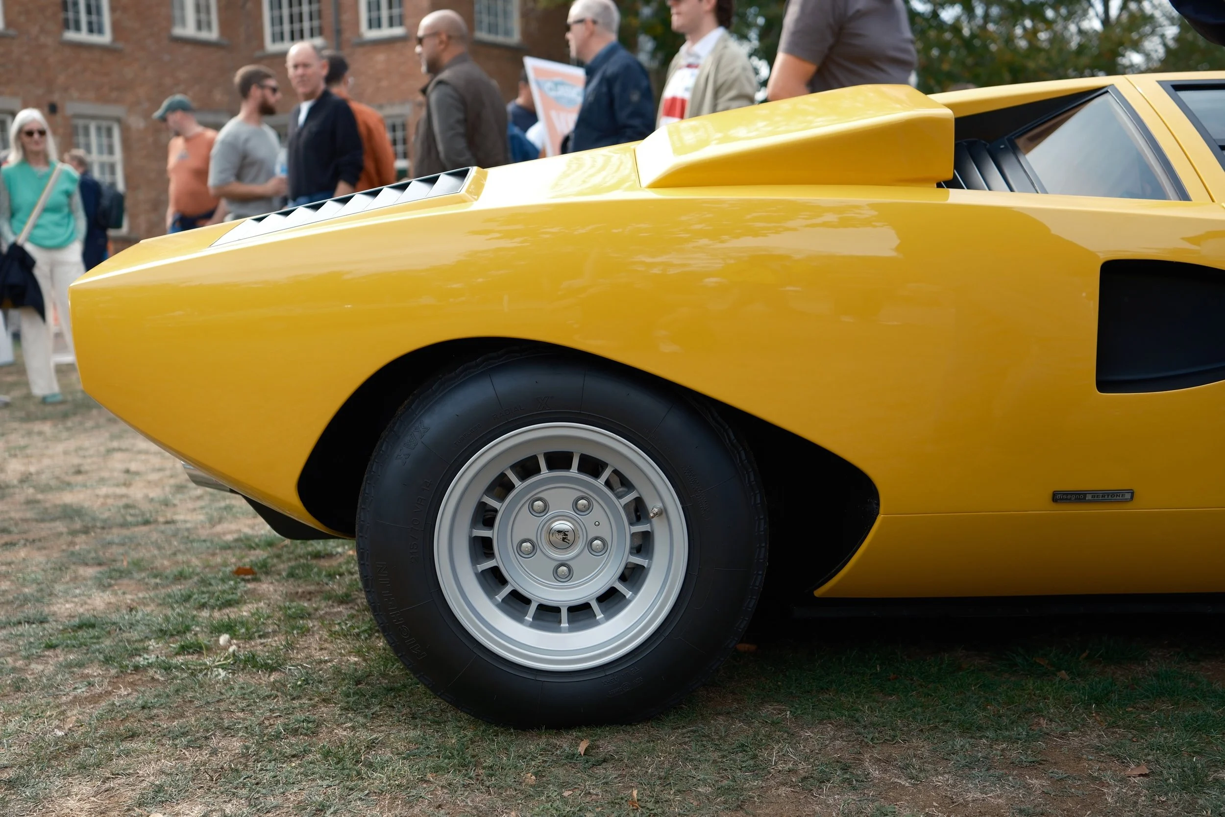 Close-up of the front portion of a yellow car with a group of people standing in the background, outdoors.