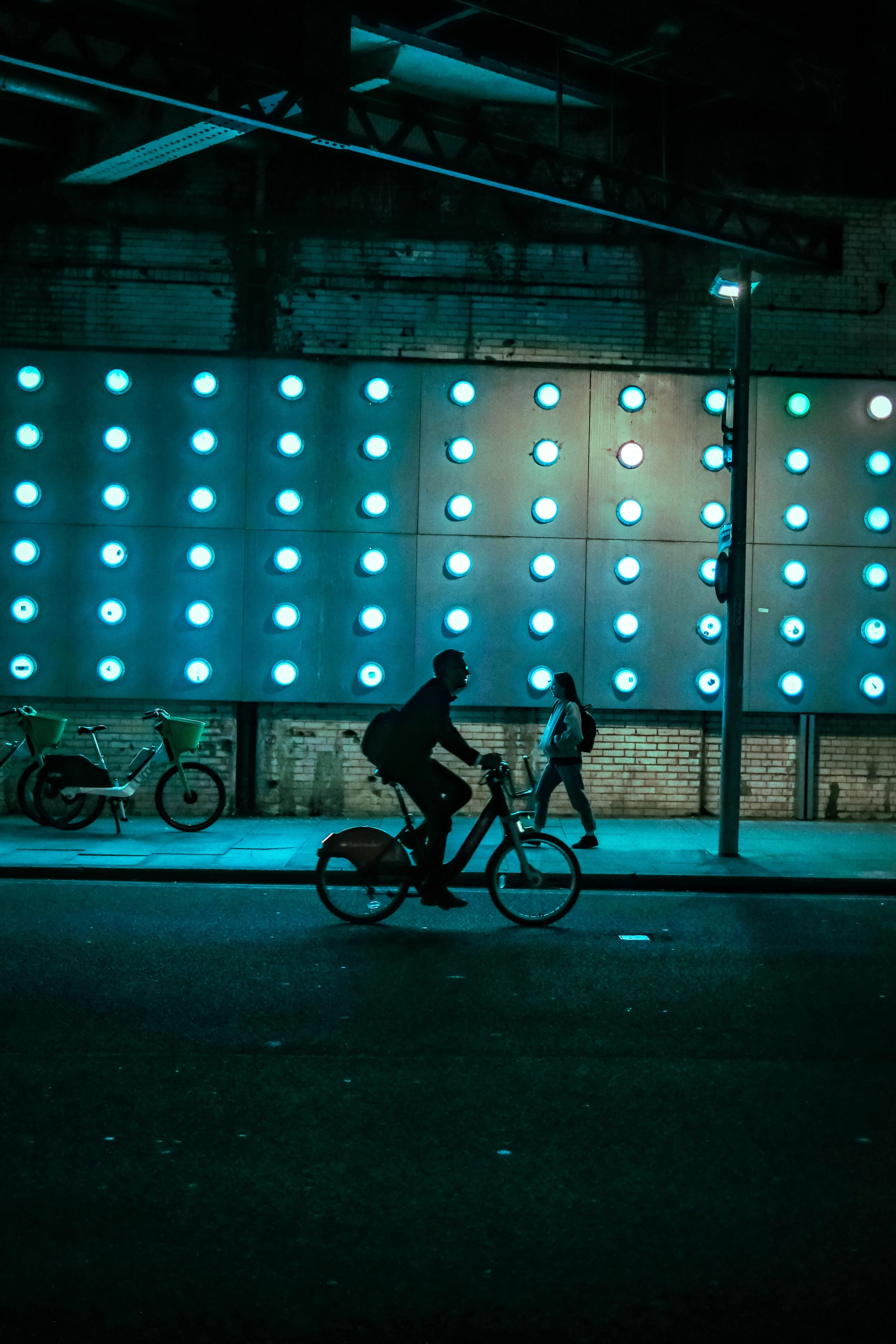 Nighttime city street scene with a decorated wall featuring a pattern of bright blue circular lights, a person riding a bicycle, and two pedestrians walking on the sidewalk, with bicycles parked along the curb.