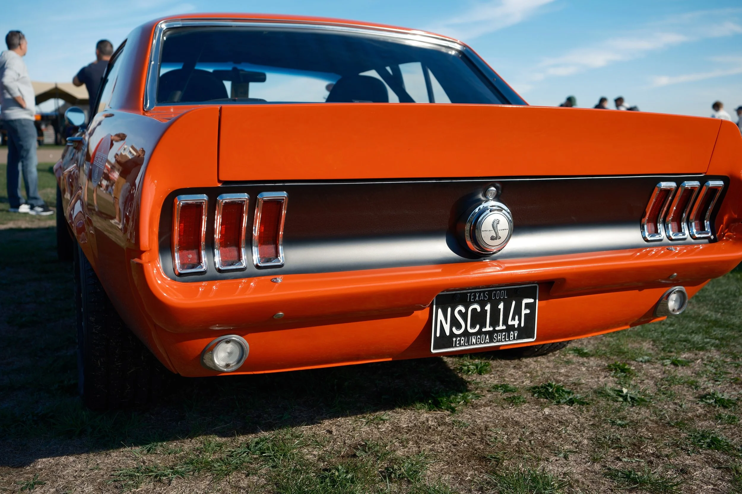 Rear view of an orange vintage Ford Shelby Mustang car parked on grass, with people in the background at an outdoor event under a blue sky.