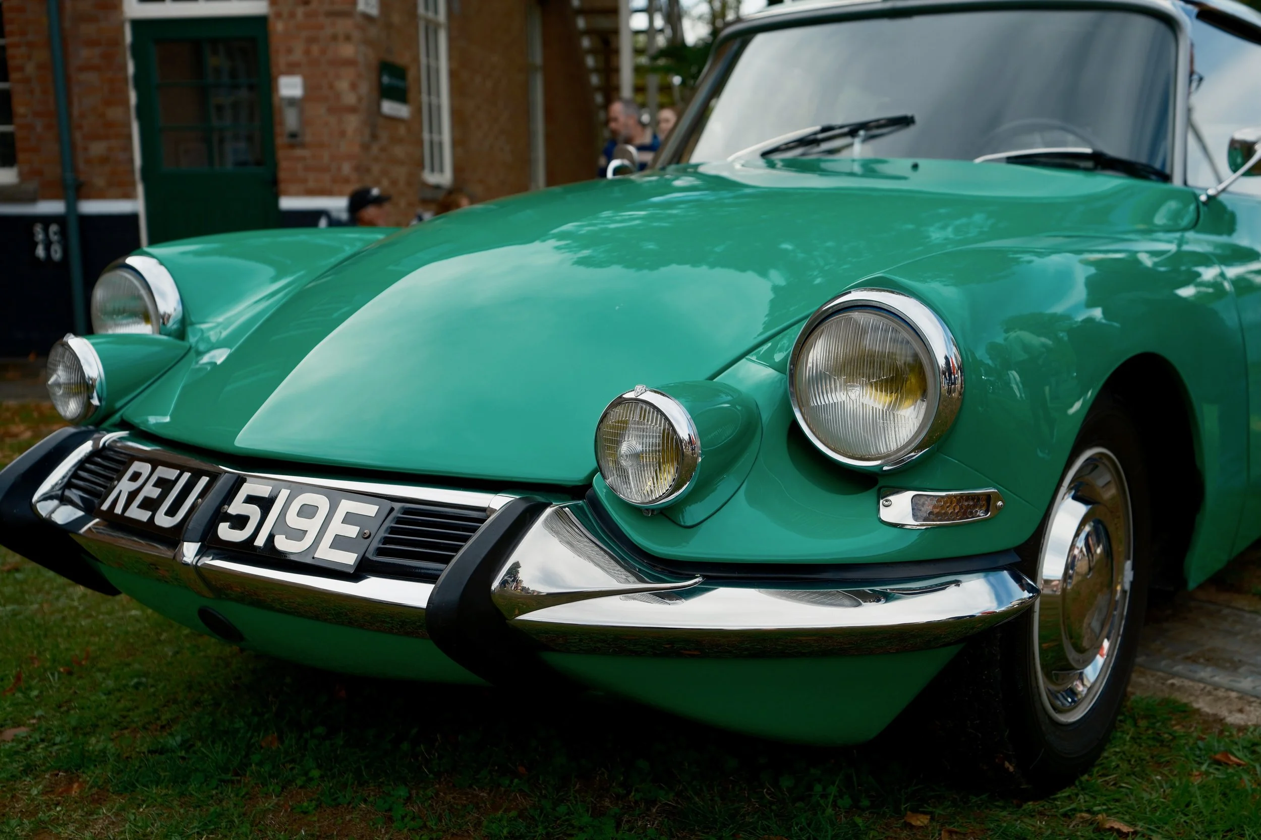 A vintage green car parked on grass in front of a brick building with people in the background.