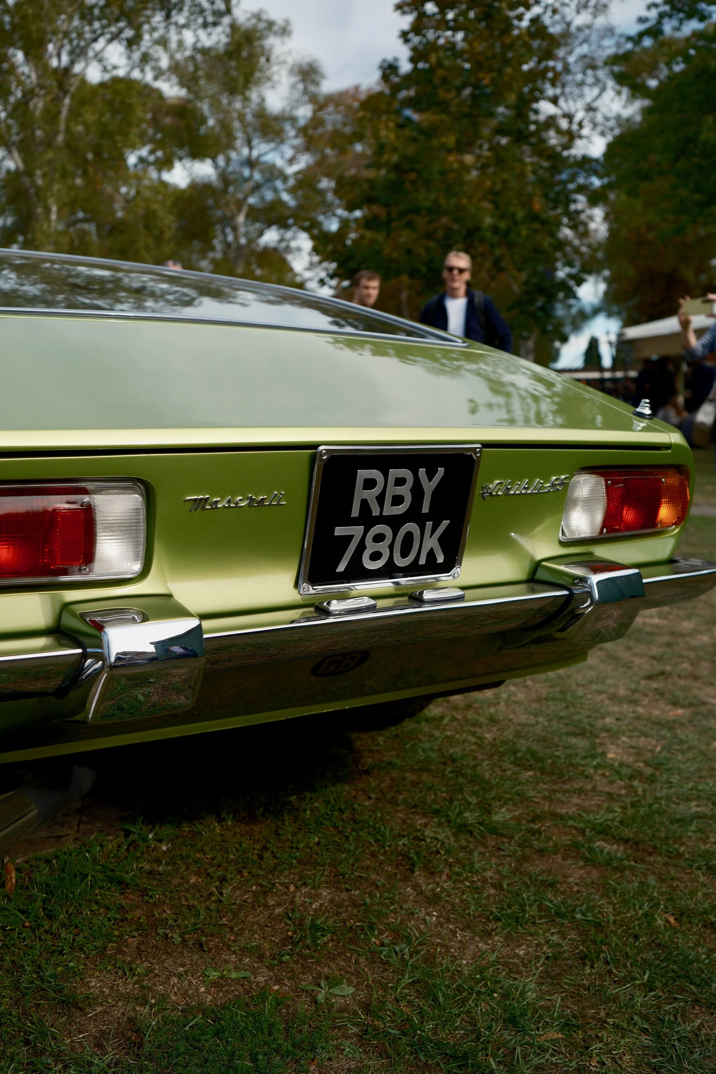 Close-up of the rear end of a lime green Maserati car with the license plate RBY 780K, parked on grass with trees and people in the background.