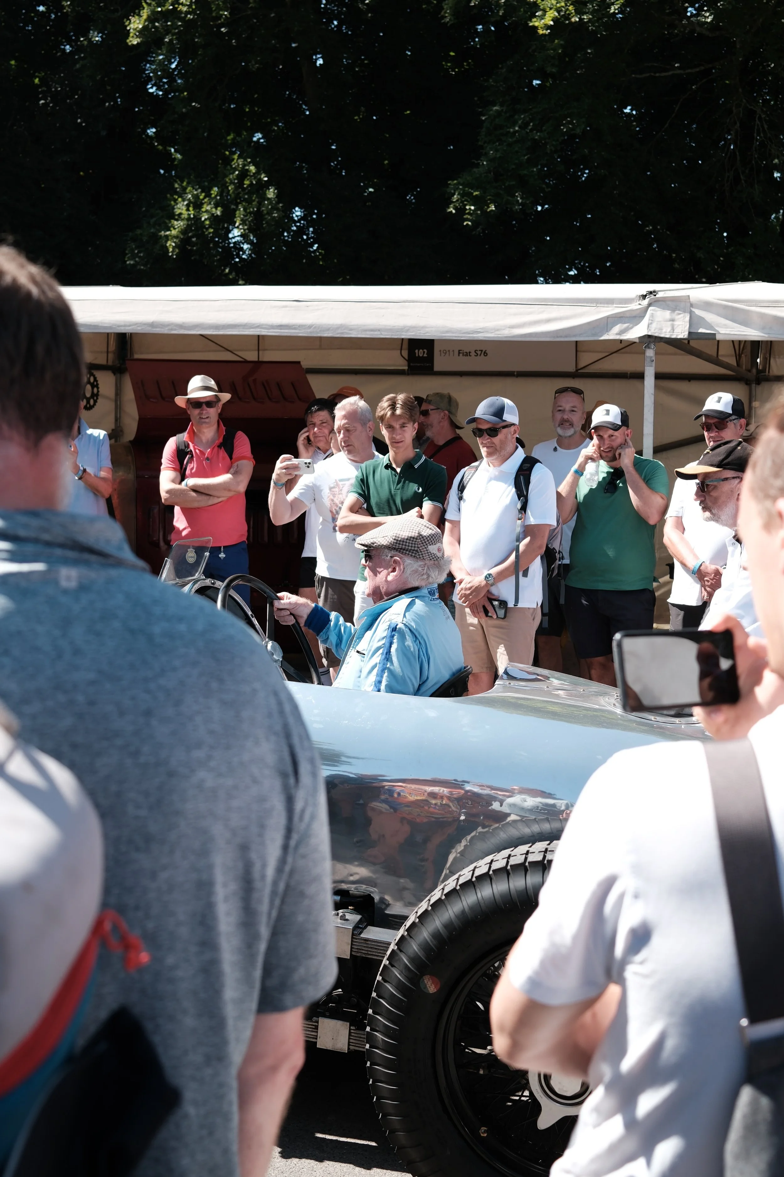 Crowd of people gathered around a shiny vintage racing car at an outdoor event, with some taking photos and others inspecting the vehicle, under a sunny sky with trees in the background.