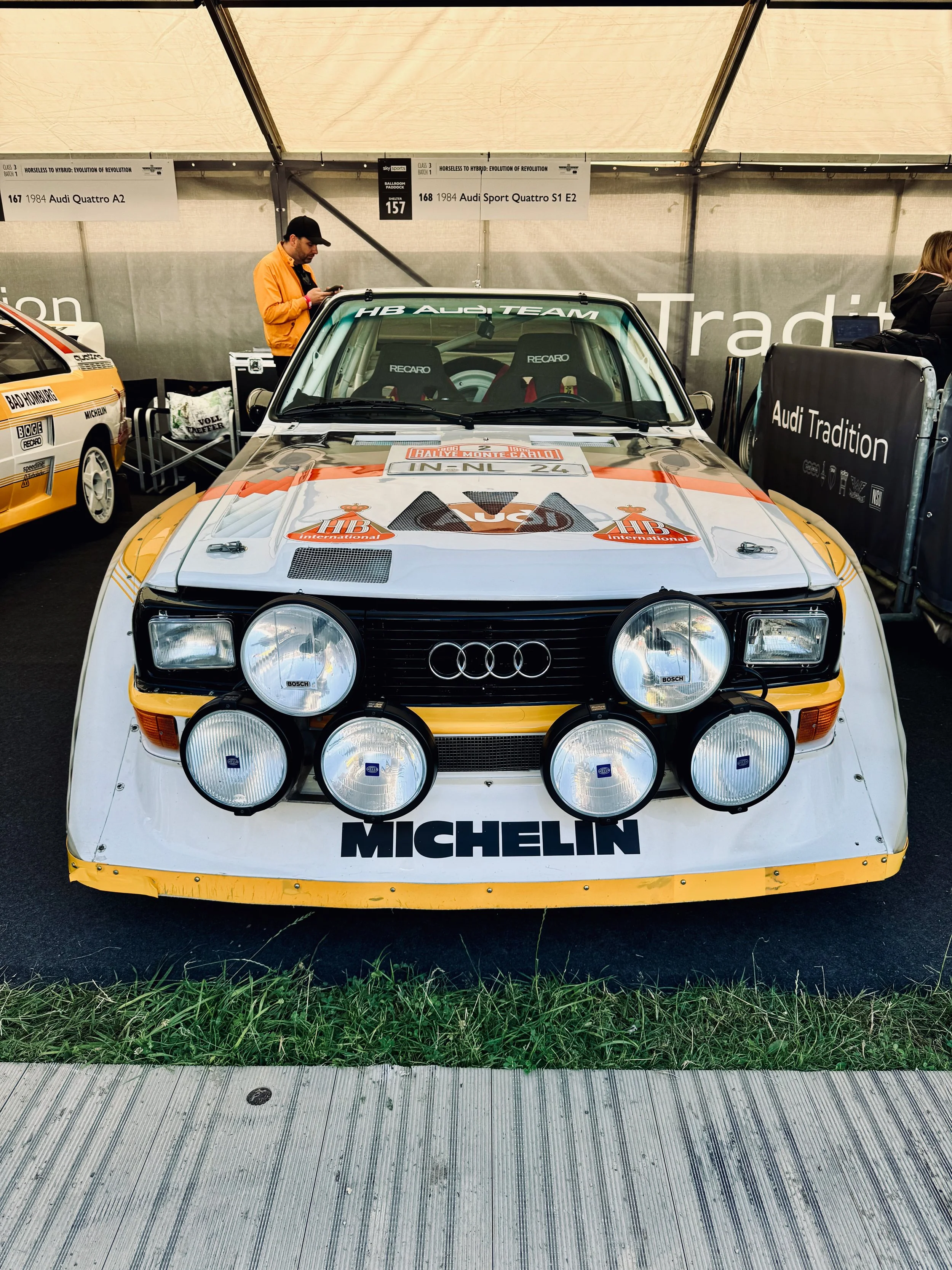 Front view of a white racing car with yellow and black accents, multiple large auxiliary lights on the front, and various sponsor logos including Michelin and Bosch, parked in a tent with a man in an orange jacket and cap looking at his phone in the 