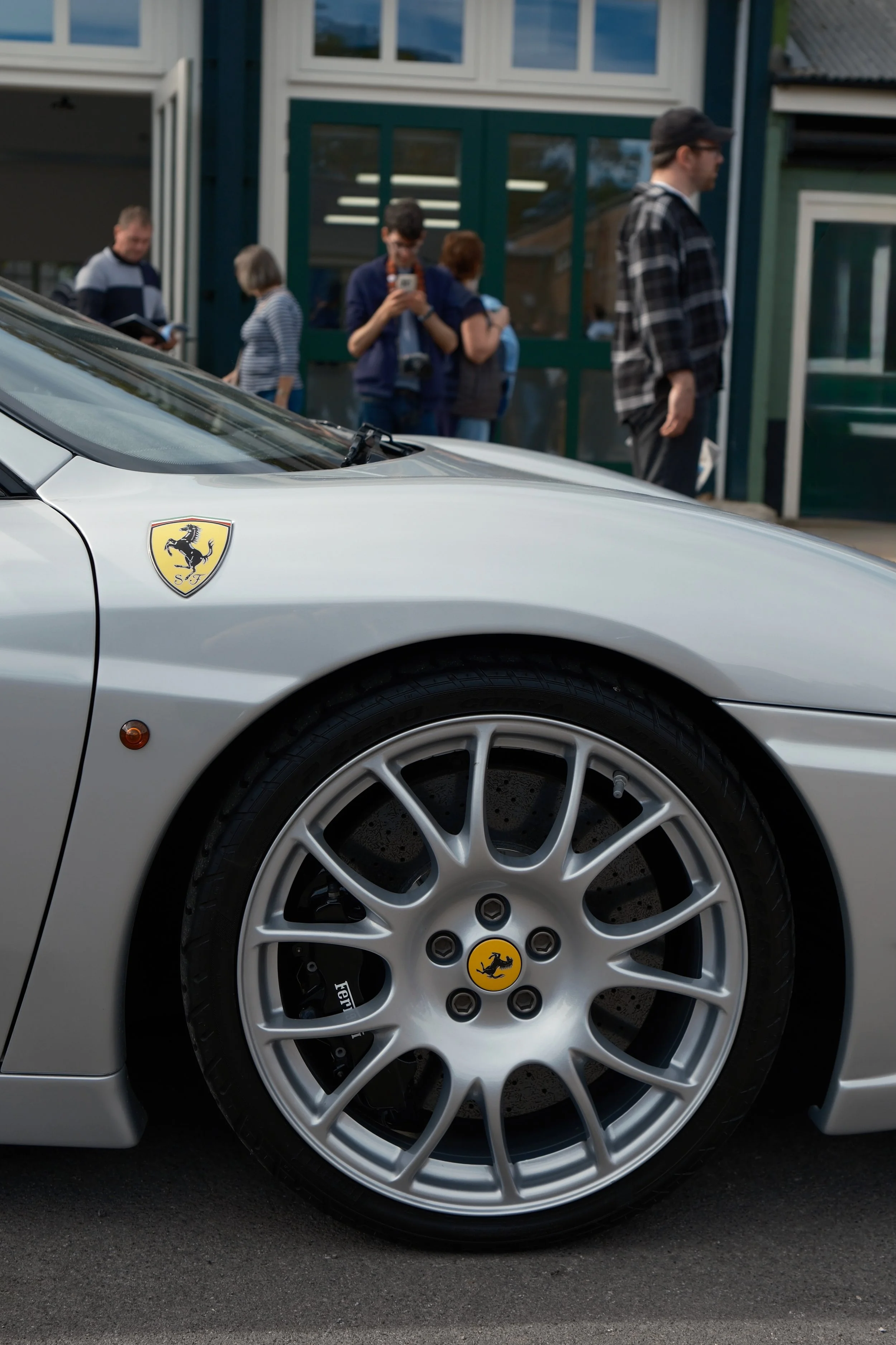 Close-up of silver Ferrari car with visible Ferrari logo on the wheel and front fender, showing a group of people in the background near a building.