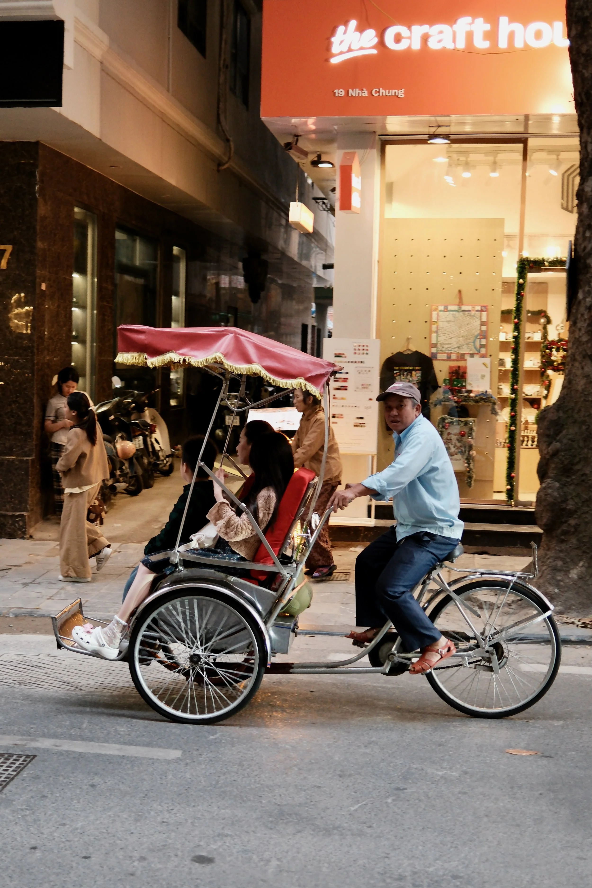A man riding a bicycle rickshaw with two young girls in the back, one girl wearing a coat and the other in a jacket, on a sidewalk in front of a store with Christmas decorations.