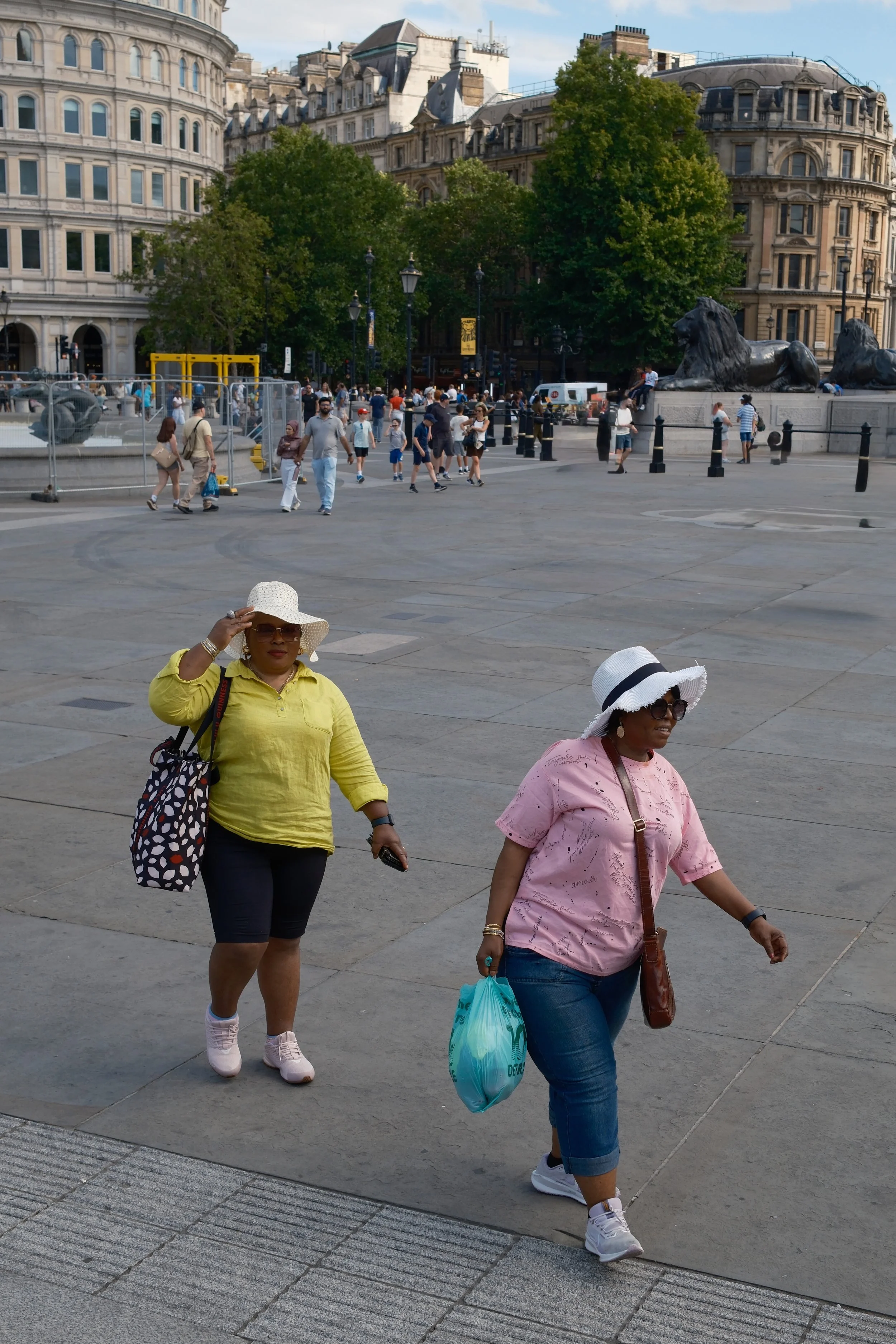 People walking in a city square with historic buildings, trees, and statues of lions in the background.