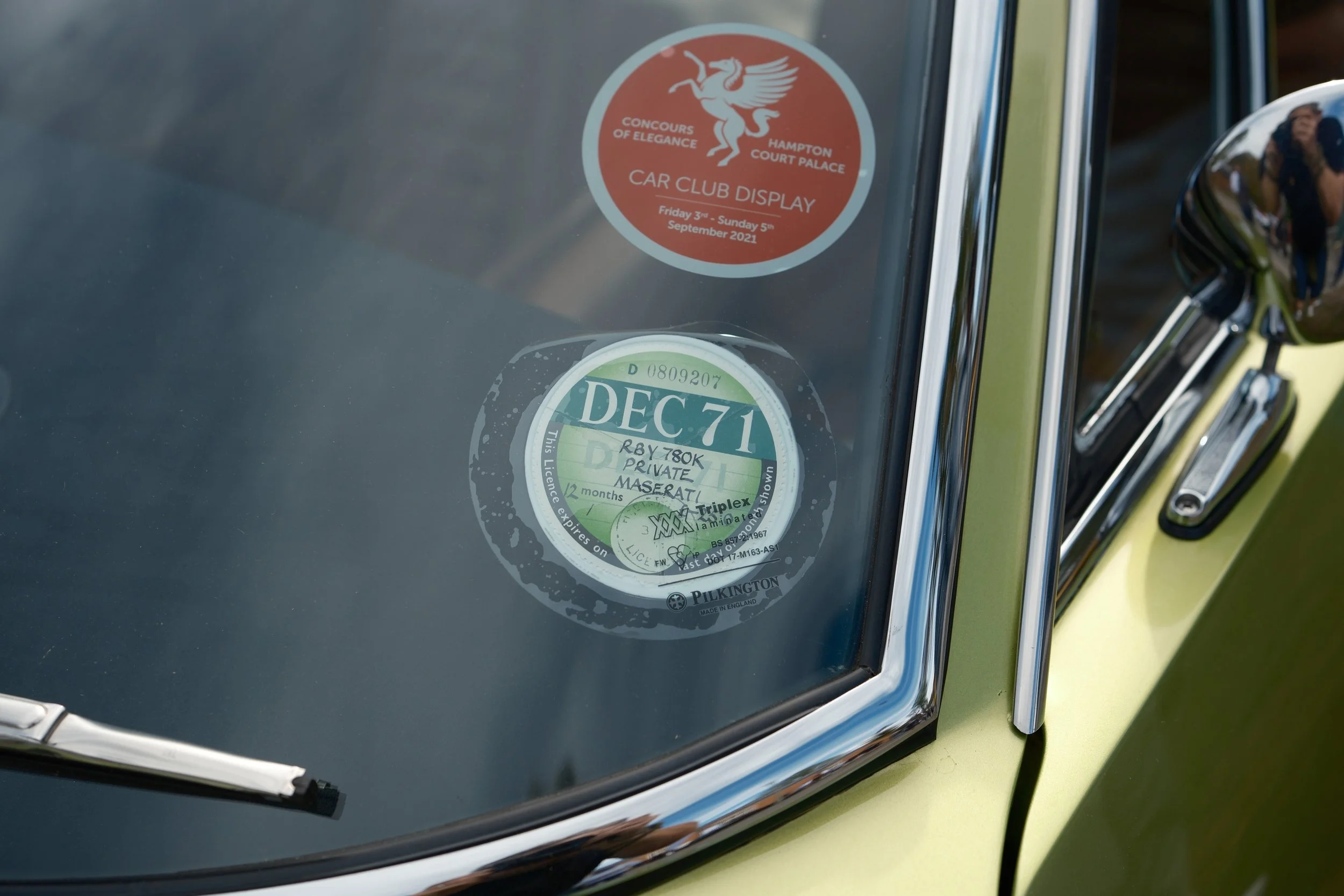 Close-up of the front windshield of a classic green car with two circular stickers, one for a car club display event and another showing a license expiration date.