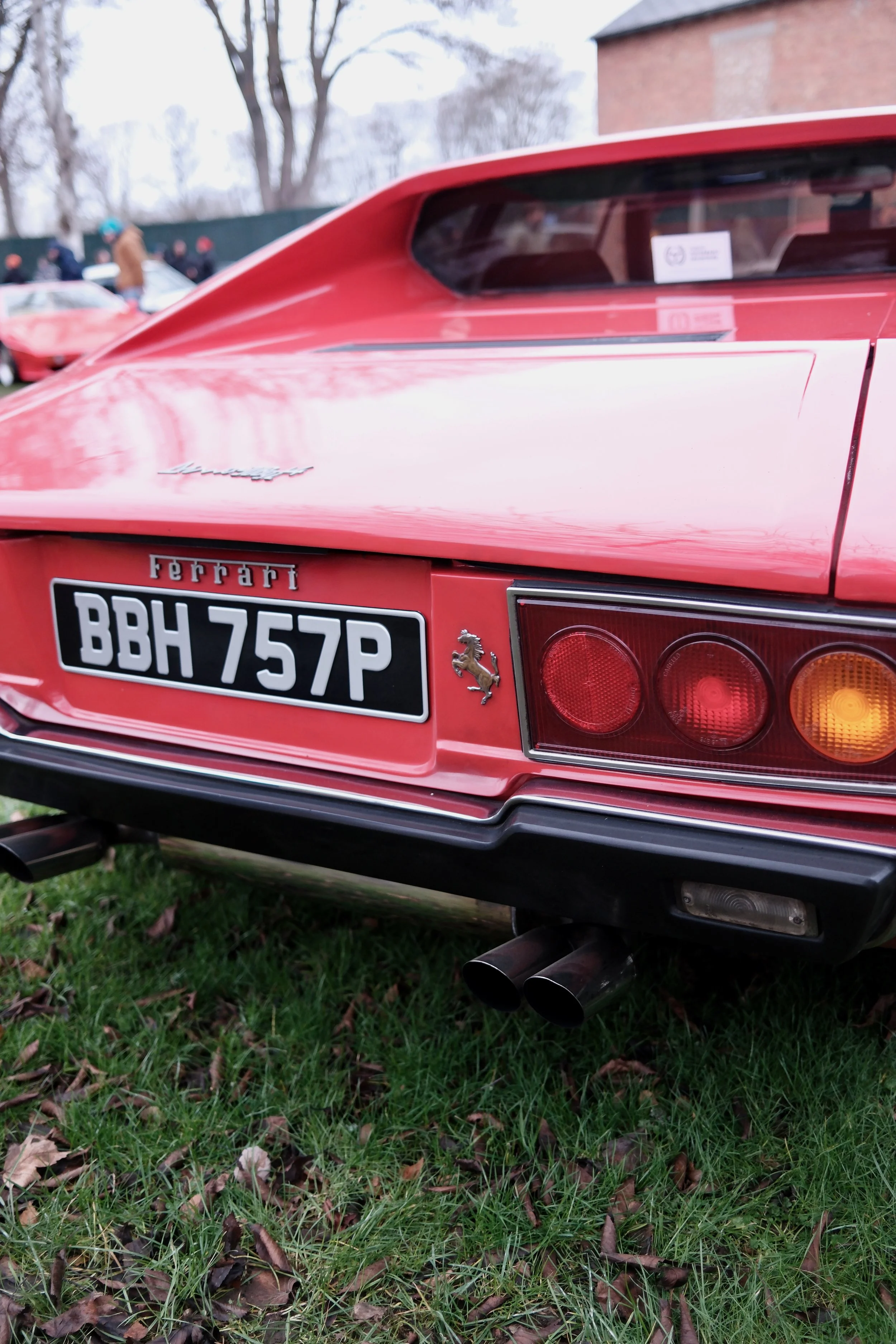 Close-up of the rear end of a red Ferrari sports car, showing the tail lights, partial rear grille, dual exhaust pipes, and a Ferrari logo badge, parked on a grassy area with fallen leaves, with other cars and people in the background.
