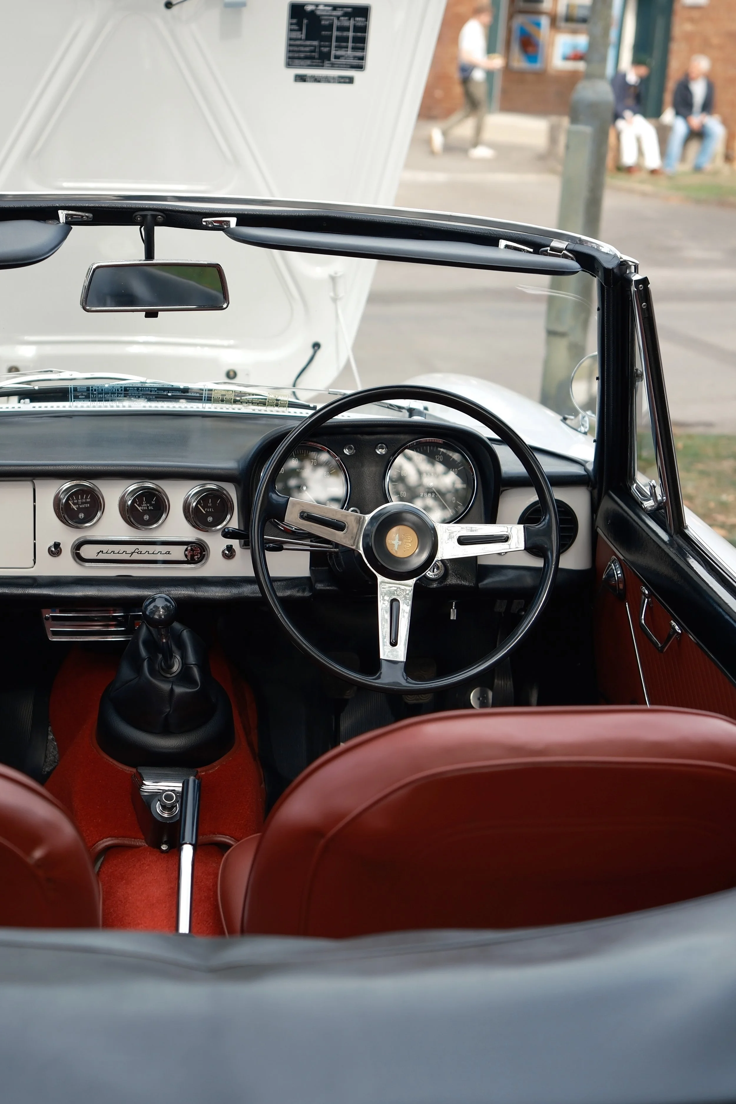 View from inside a classic car showing the steering wheel, dashboard, and red seats, with the open hood and a street scene with people sitting and walking in the background.