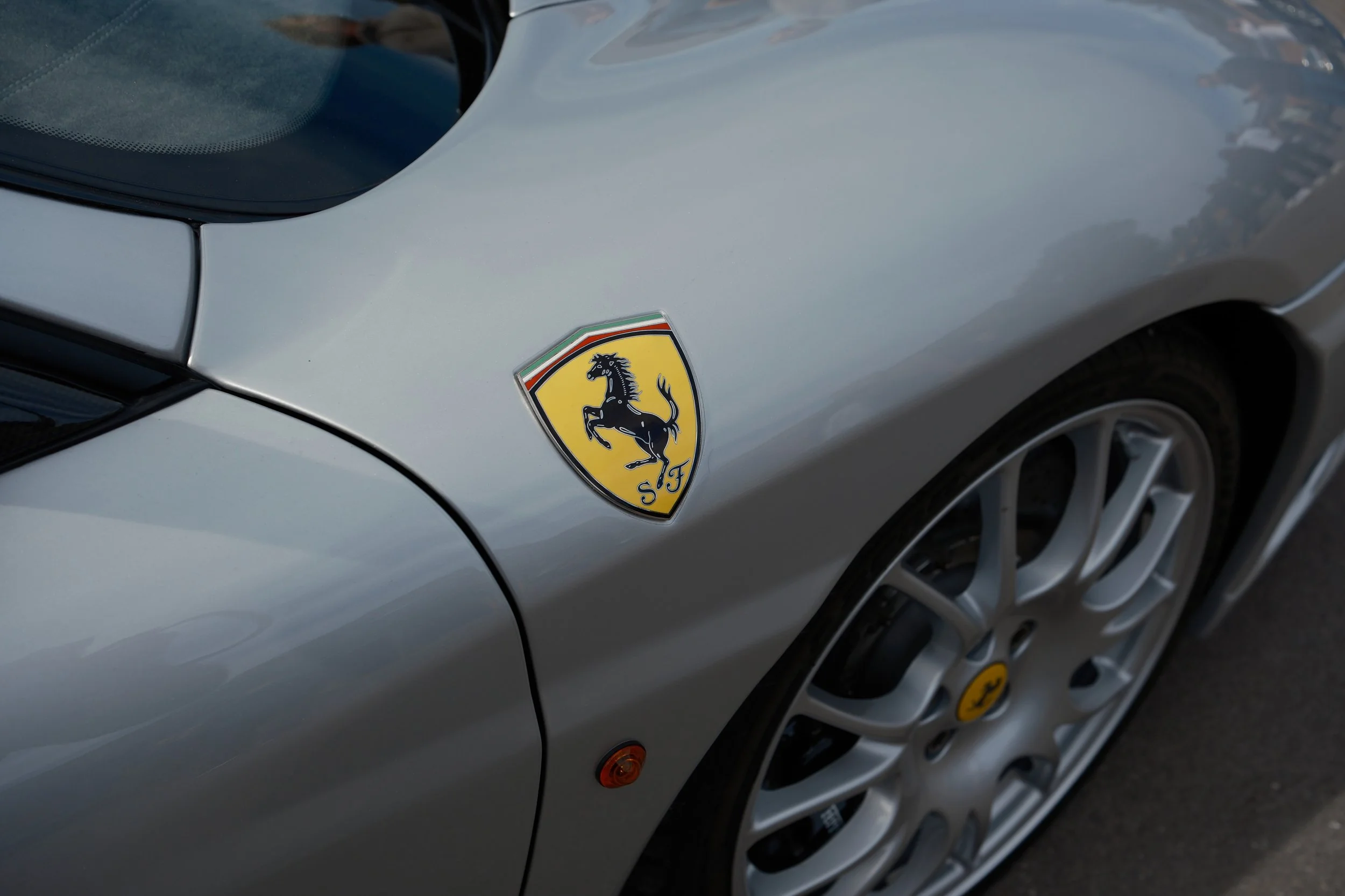 Close-up of a silver Ferrari car's front fender, showing the Ferrari logo with a black horse on a yellow shield and a part of the wheel with a silver rim.