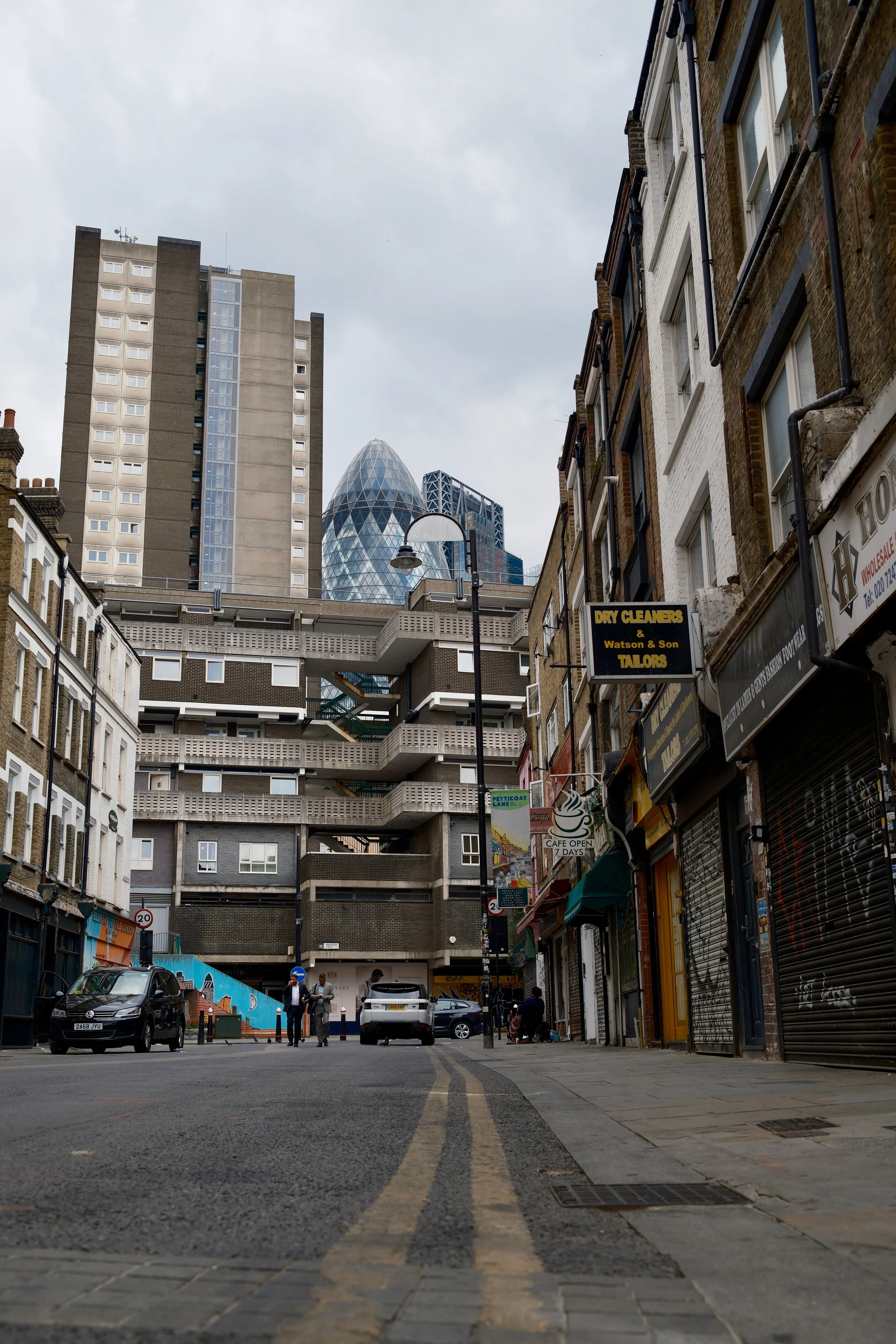 A city street view with parked cars, pedestrians, and tall modern buildings, including the Gherkin, in London's financial district under cloudy skies.