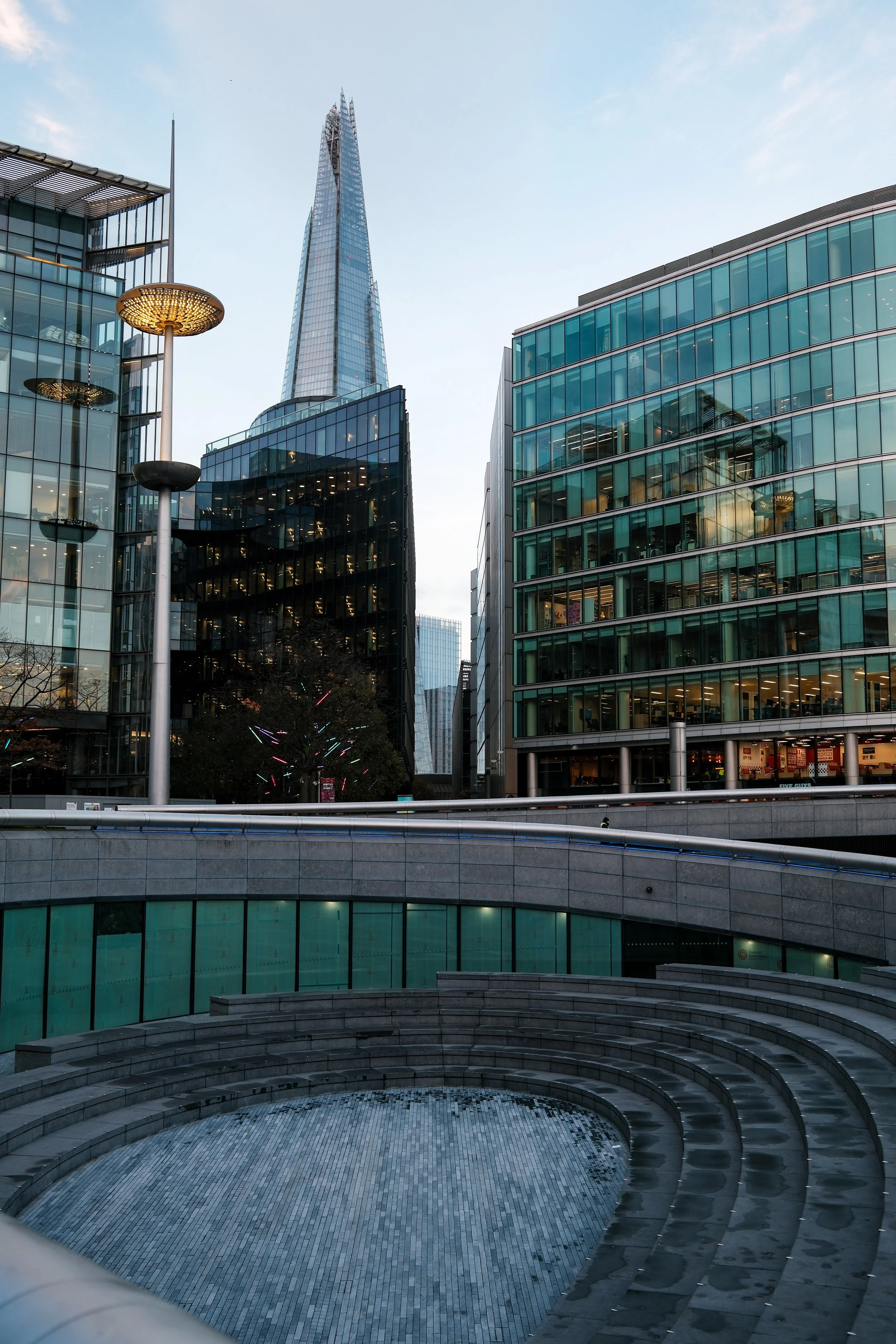 A cityscape featuring modern glass buildings with a tall, tapering skyscraper in the background. There are pathway steps in the foreground, some streetlights, and a tree with colorful lights. The sky is mostly clear with a few clouds.