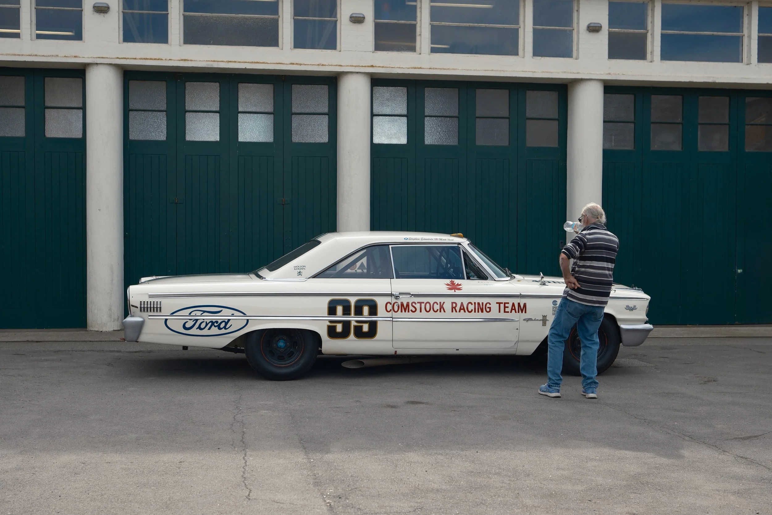 A man with gray hair drinking water from a plastic bottle next to a vintage white Ford race car with number 99 and racing decals, parked on an empty street with a large green door building in the background.