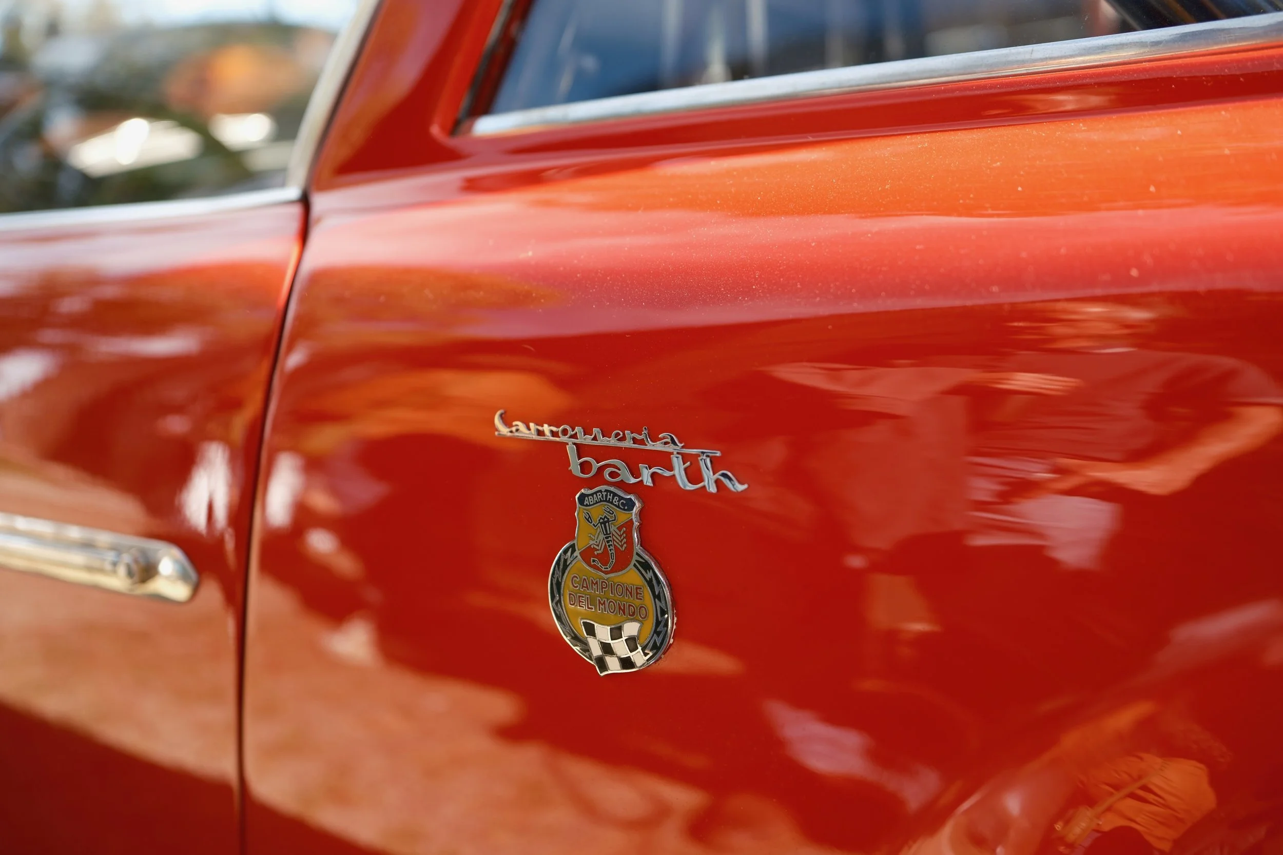 Close-up of a red vintage car's side panel with a badge that reads 'Carrozzeria Bart,' featuring a shield emblem and an Italian flag. The car's shiny paint reflects the surroundings.