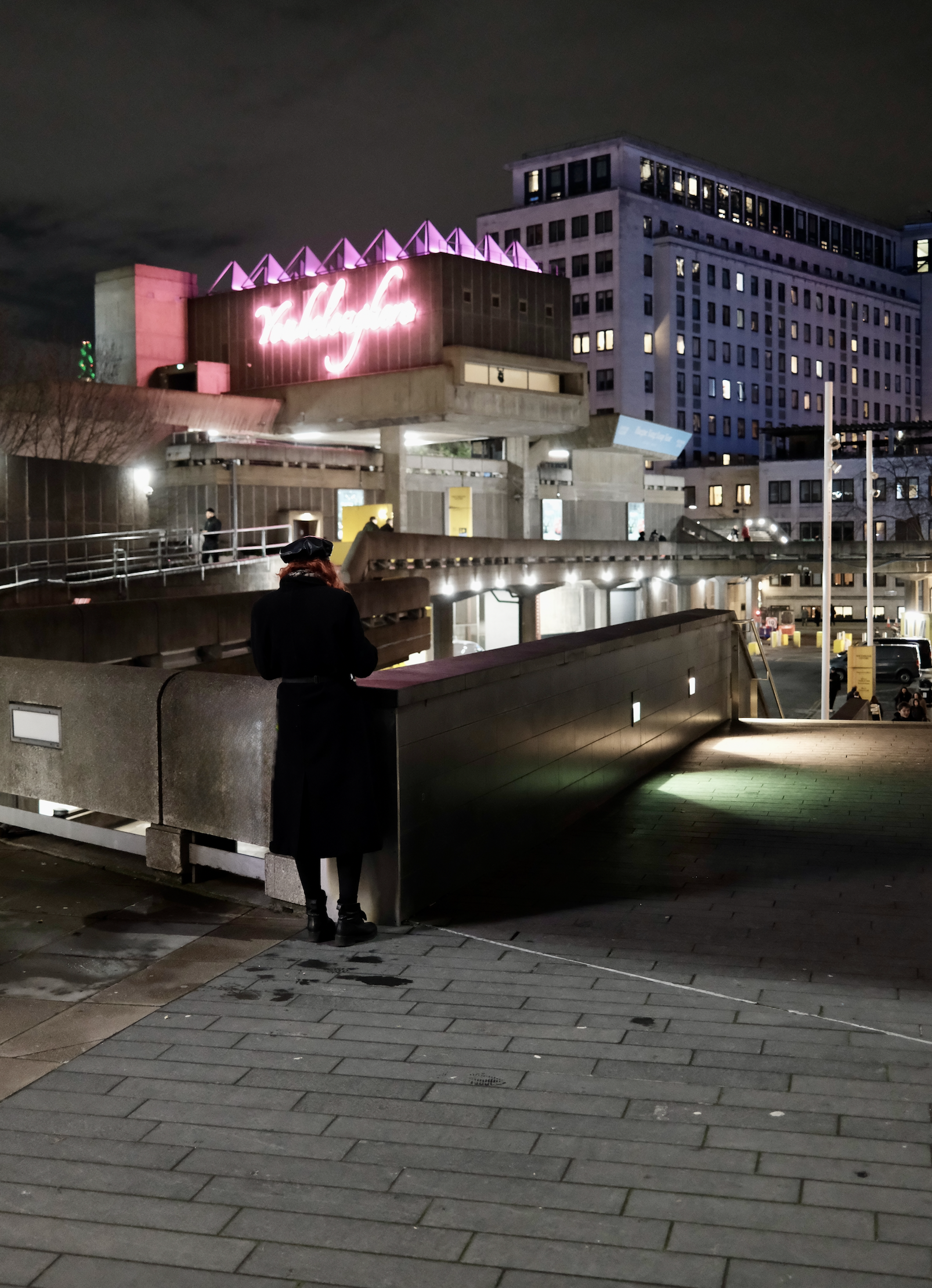 Nighttime city scene with a person in black coat and hat, standing on a paved walkway near a concrete railing, with a large building and neon sign in the background.