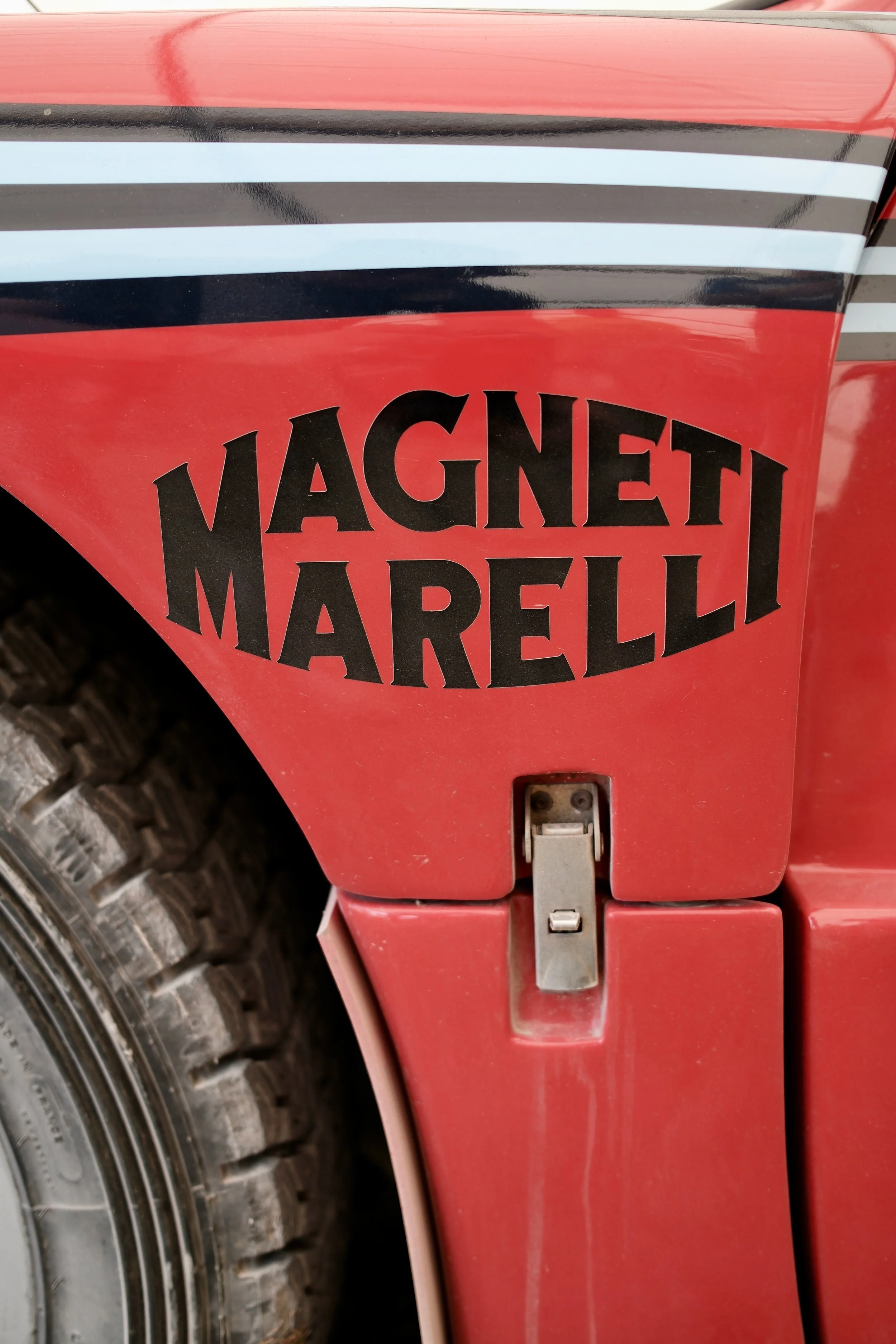 Close-up of a red vintage tractor with black and white stripes and a sticker that reads 'Magneti Marelli' on the side. Part of a large tire is visible in the lower left corner.