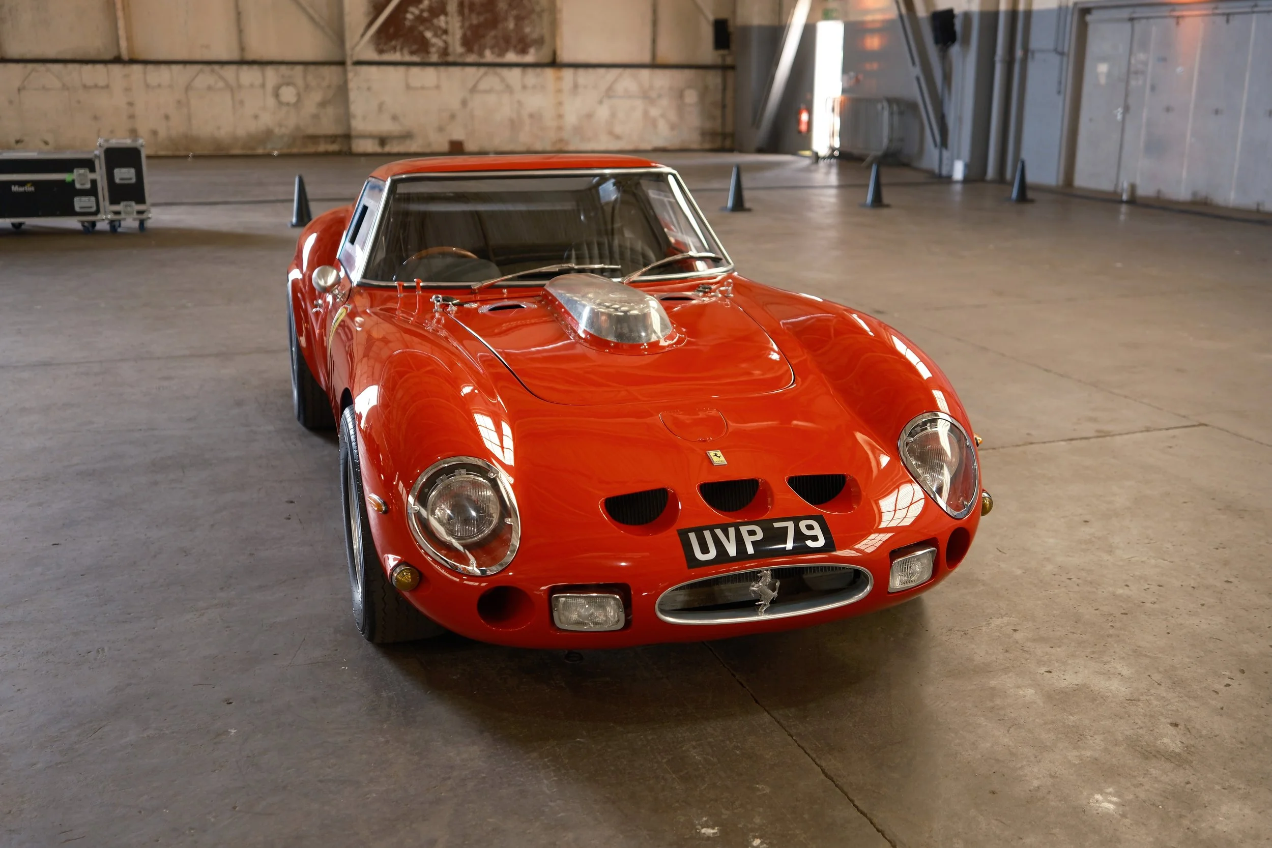 A red vintage Ferrari sports car with a silver hood scoop, parked inside a large industrial warehouse with concrete floors and brick walls.