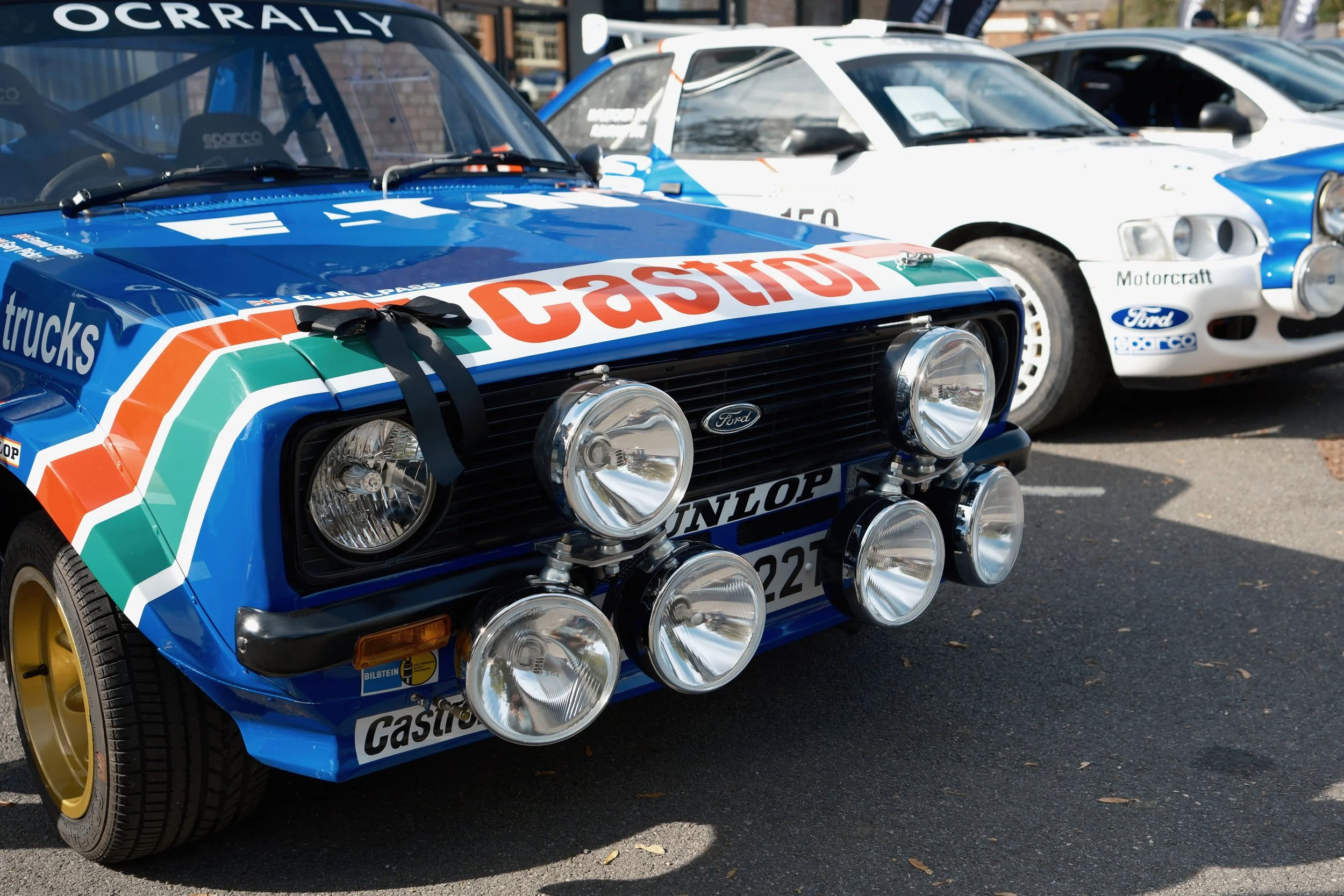 A classic rally car adorned with Castrol livery and multiple auxiliary lights parked alongside other vintage cars.