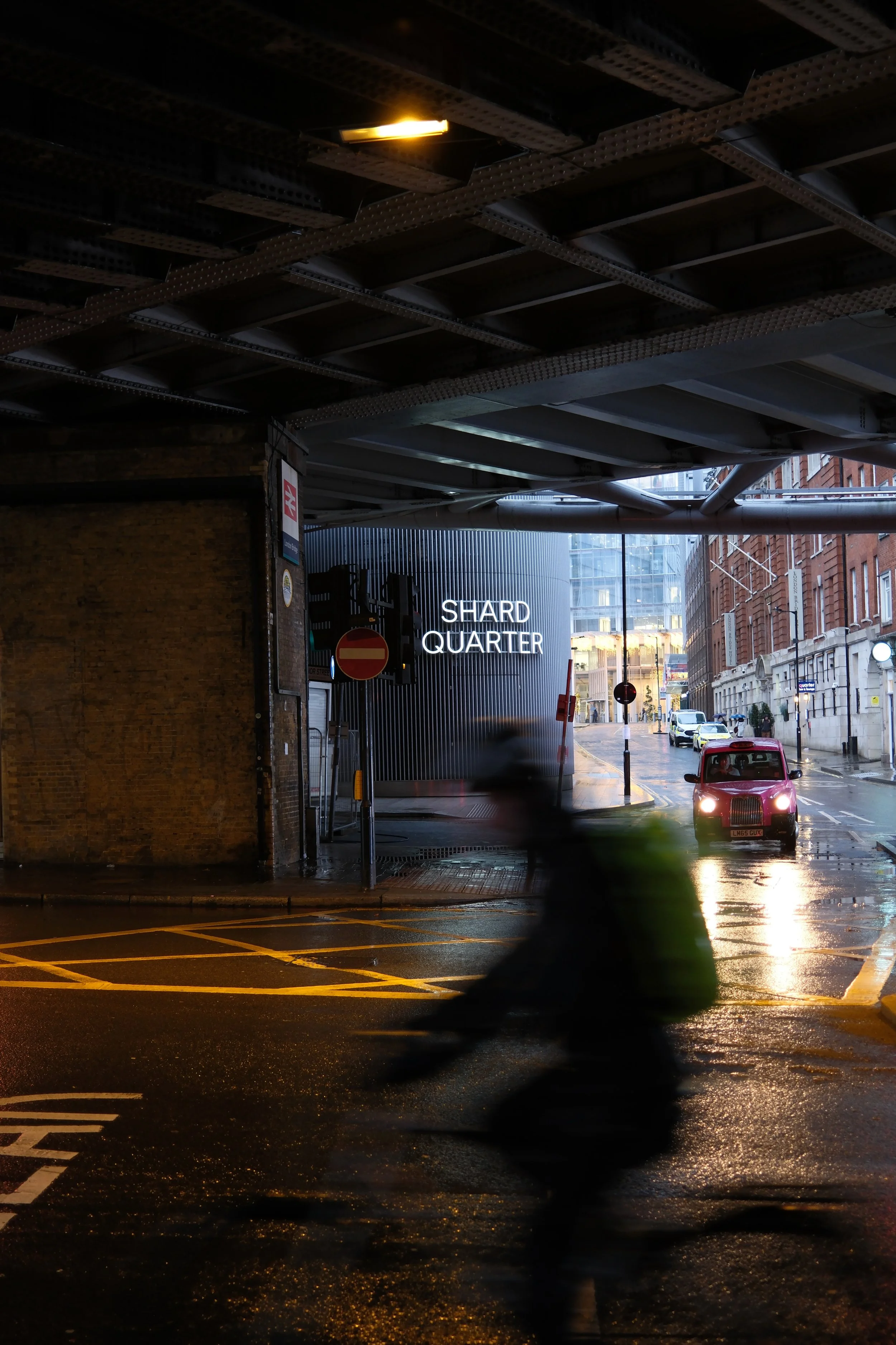 Underpass with wet street, blurred passing vehicle, illuminated street signs, buildings in the background, and a sign indicating 'Shard Quarter' in London.