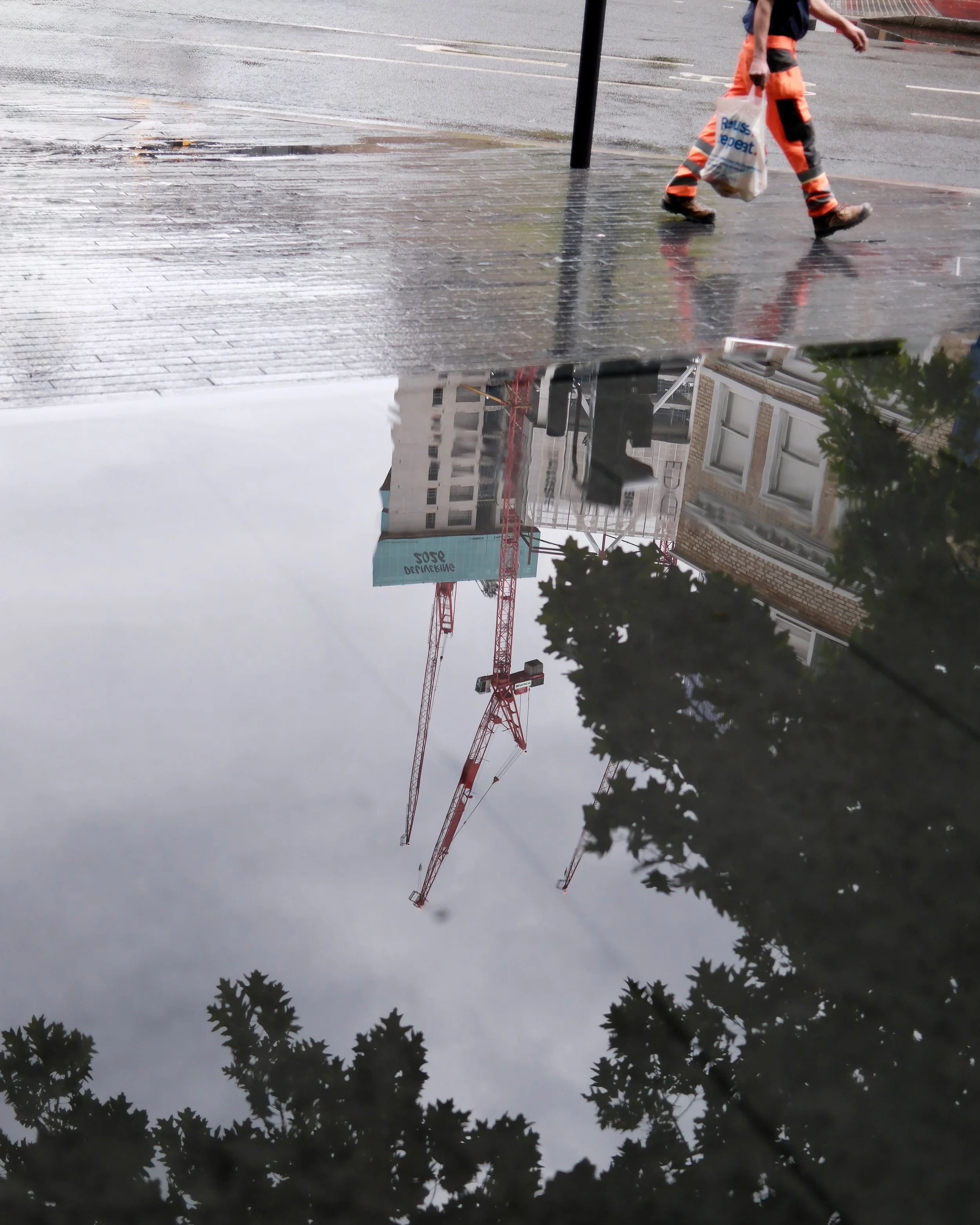 Reflected image of a street scene showing a construction crane, buildings, and trees on a wet surface. A person with orange and black work pants is walking with a bag in their hand.