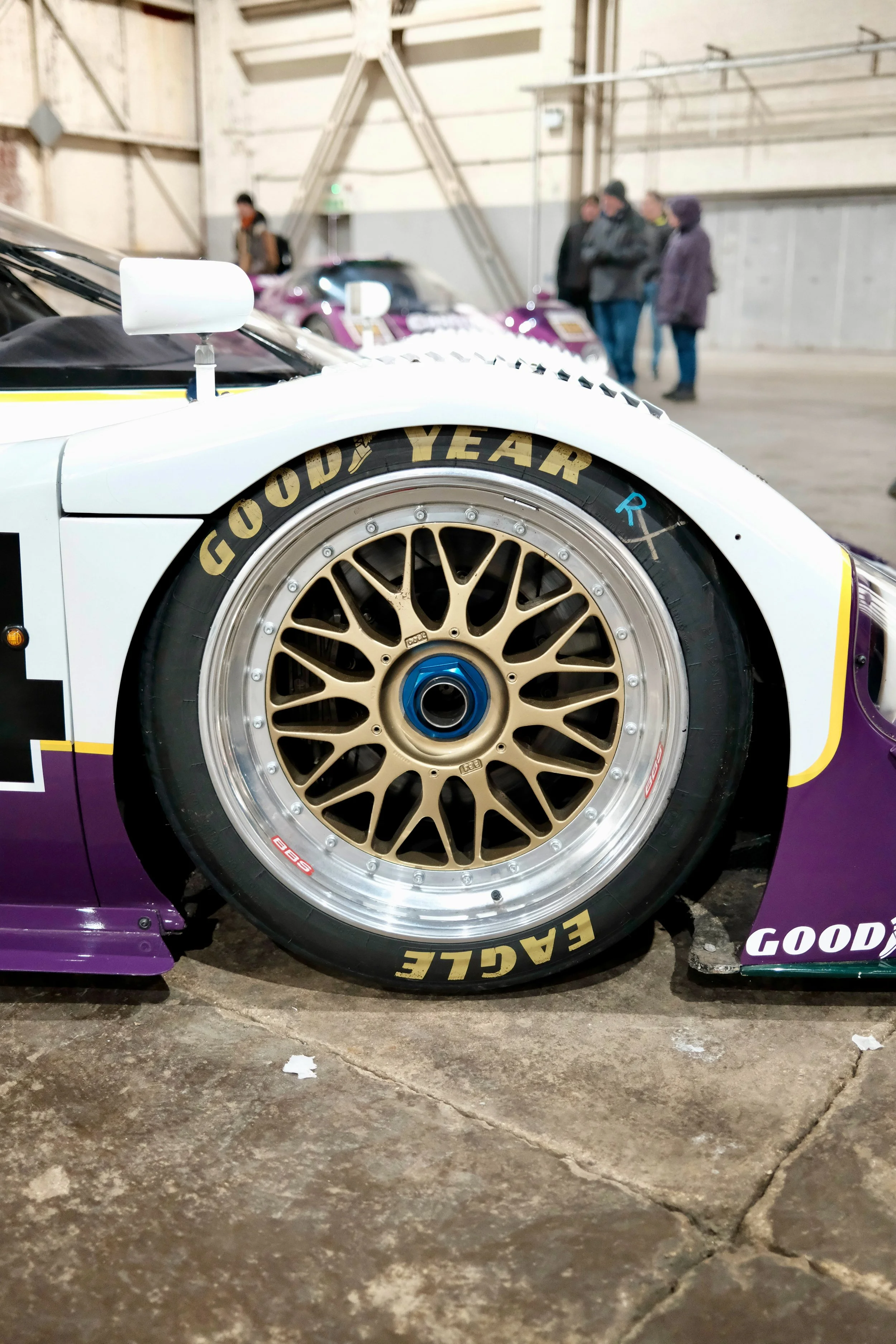 Close-up of a race car's front wheel and part of the body, with a group of people in the background inside a large industrial building or garage.