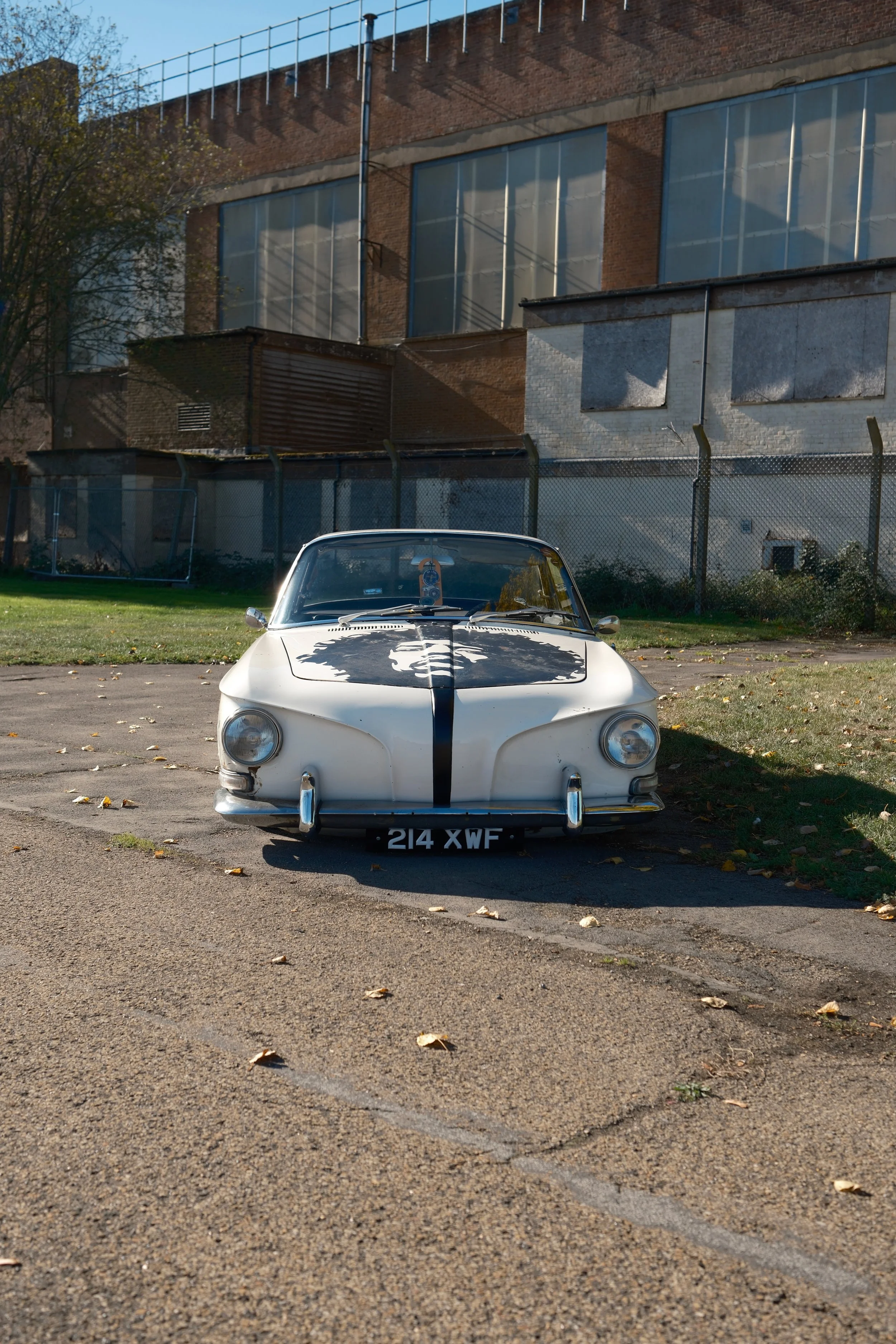 A vintage white sports car parked on an asphalt lot, with a brick building and chain-link fence in the background, under a clear blue sky.