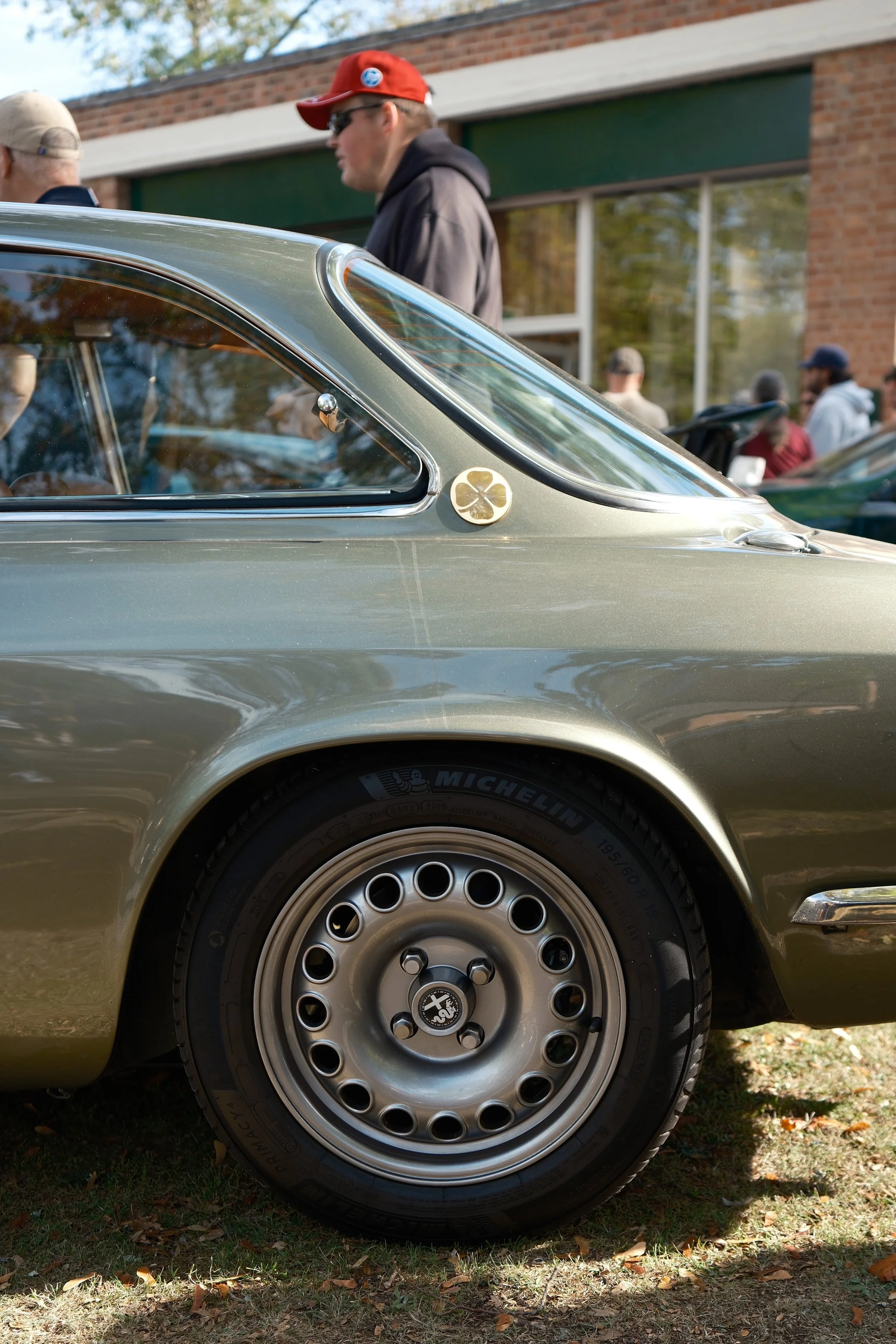 Close-up of a classic silver car's front left side, showing the wheel and part of the door, with a four-leaf clover emblem on the car's body.