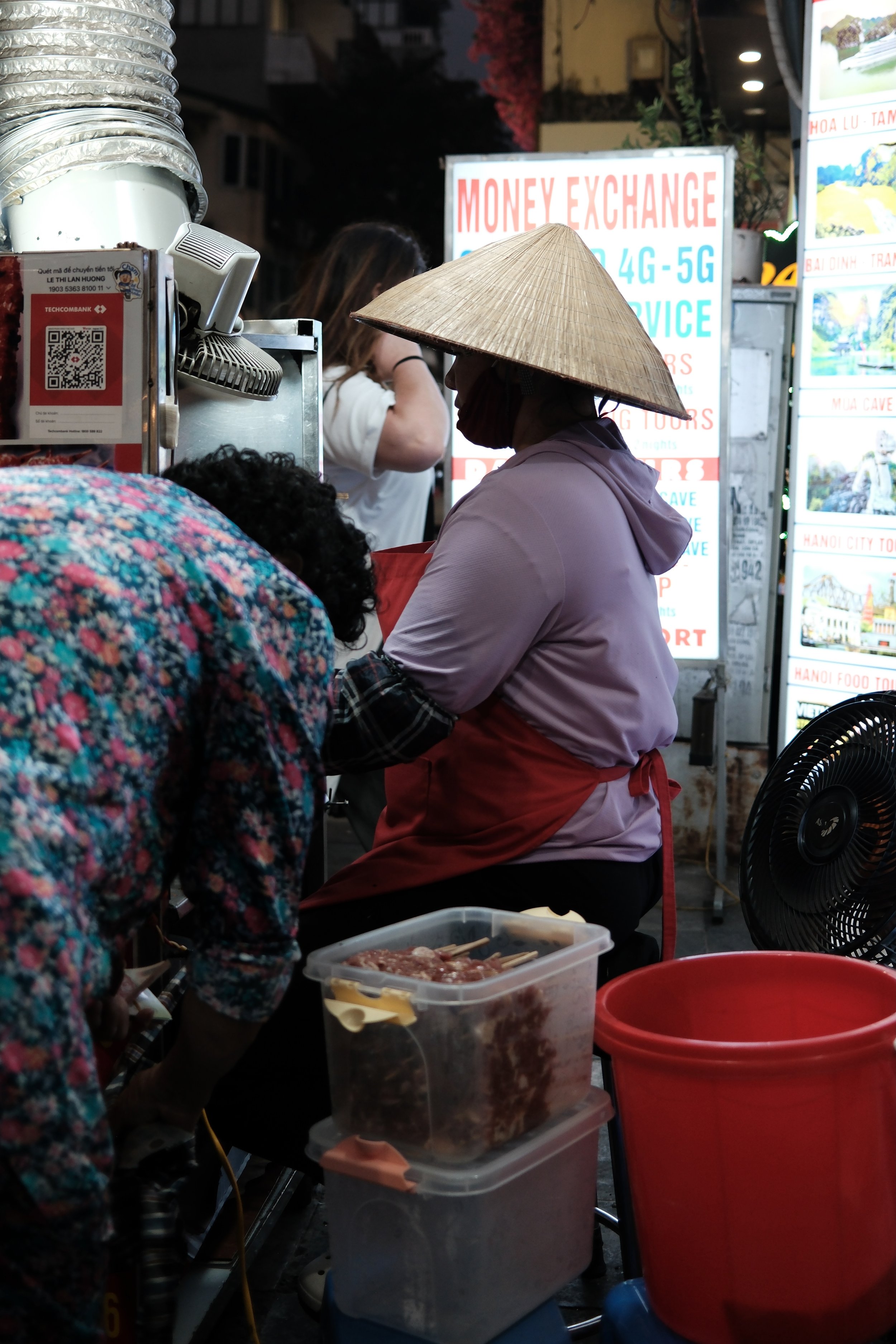 A street scene at night showing a woman wearing a traditional Vietnamese conical hat working at a food stall, with containers of ingredients, a fan, and signs advertising currency exchange and travel services in the background.