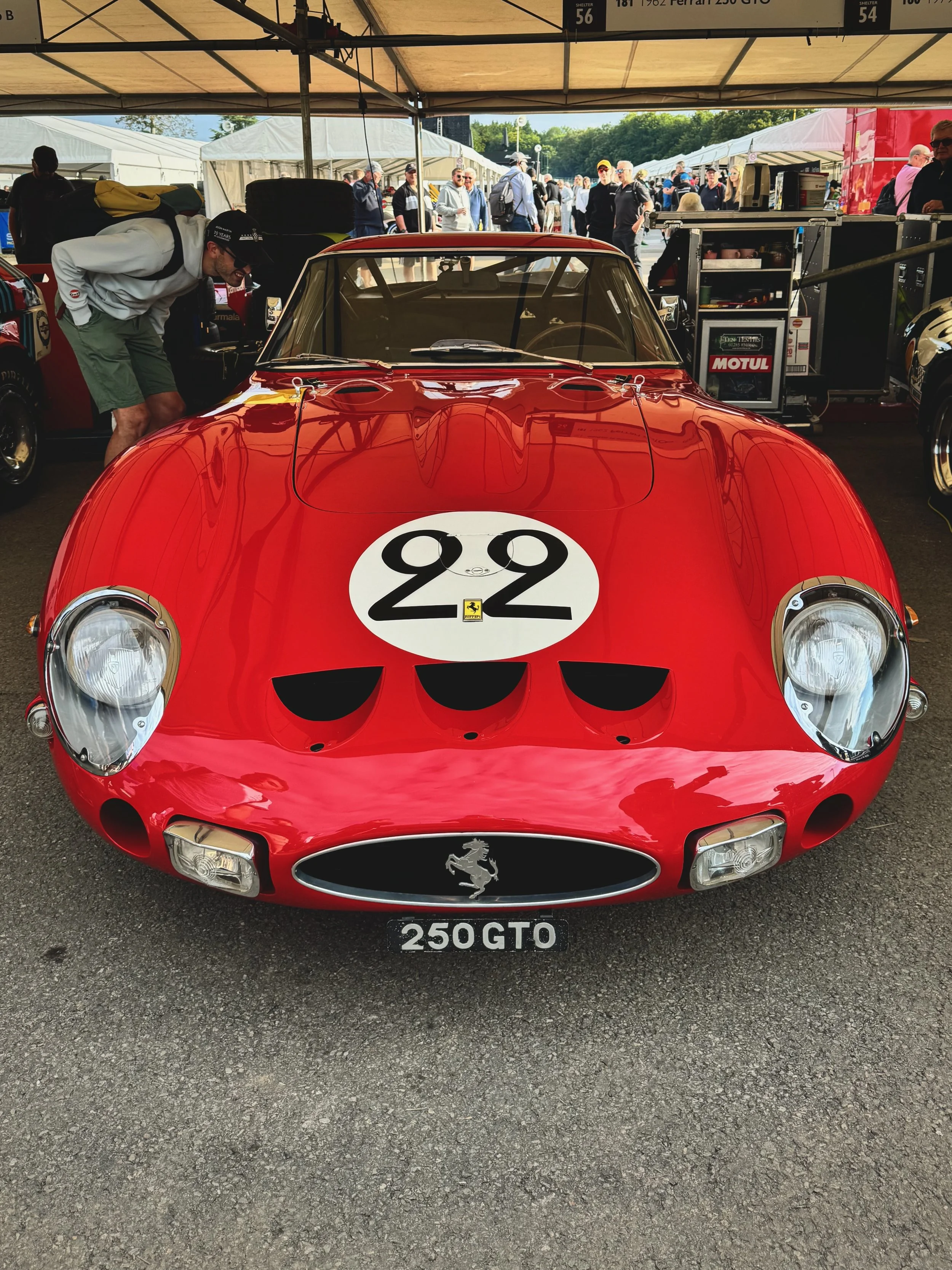 A red Ferrari race car with the number 22 on the hood, parked under a tent at a racing event. The front of the car features the Ferrari logo and the license plate reads 250GTO. Several people are around, some examining the car and others in the backg