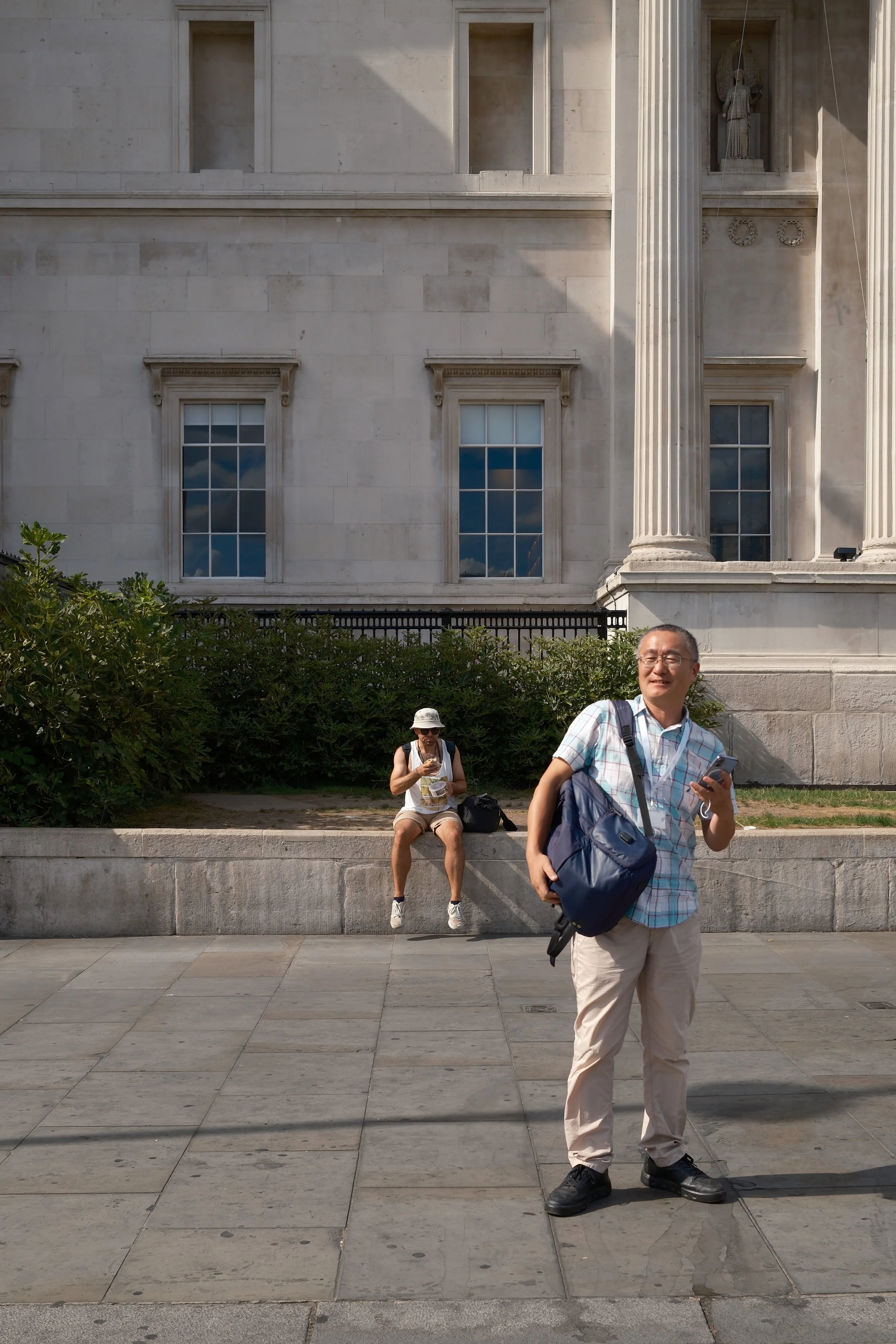 Two tourists are in front of the historic building, one standing and smiling while holding a phone, the other sitting on a ledge wearing a white hat and checking a map or phone, with a green bush and an old stone building with columns and statues beh