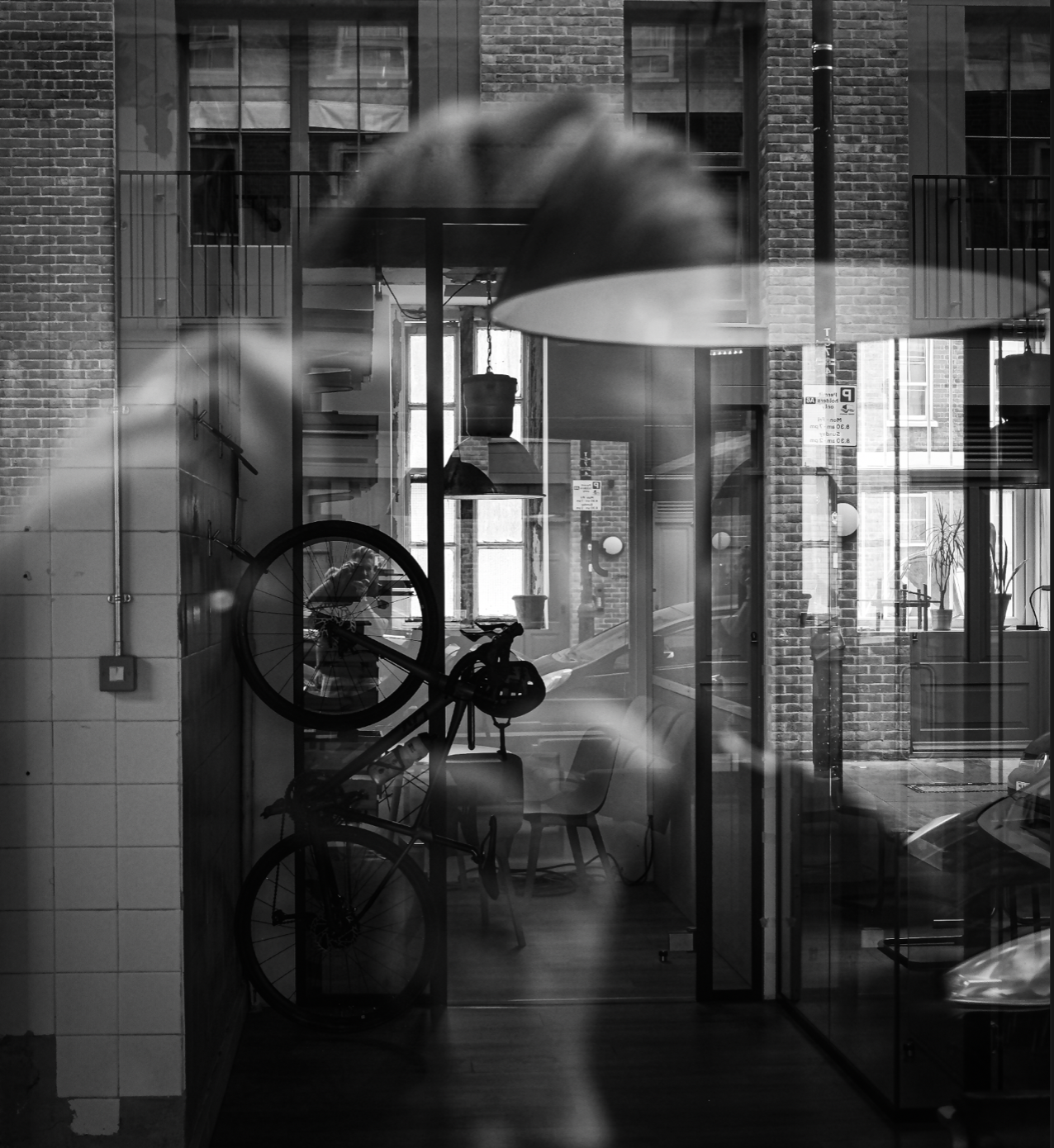 Black and white photo of a cafe or shop interior with a bicycle and a person with an umbrella outside, seen through glass windows.