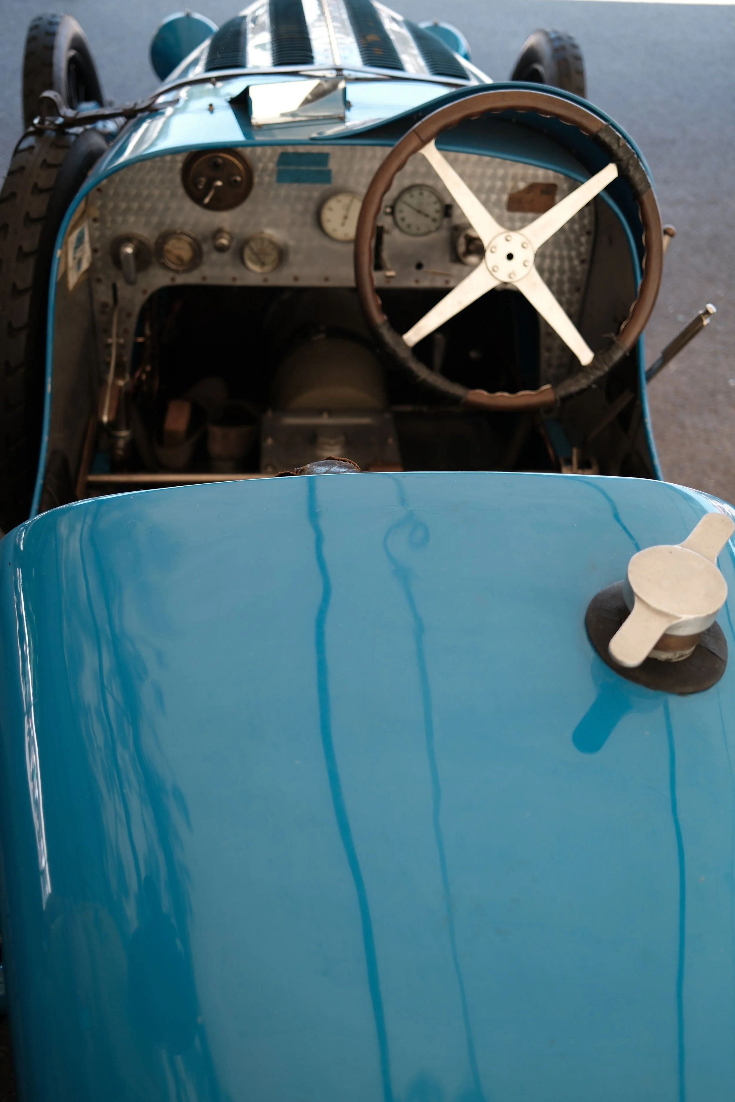 Interior view of a vintage race car dashboard and steering wheel, with gauges, switches, and a blue body exterior visible.