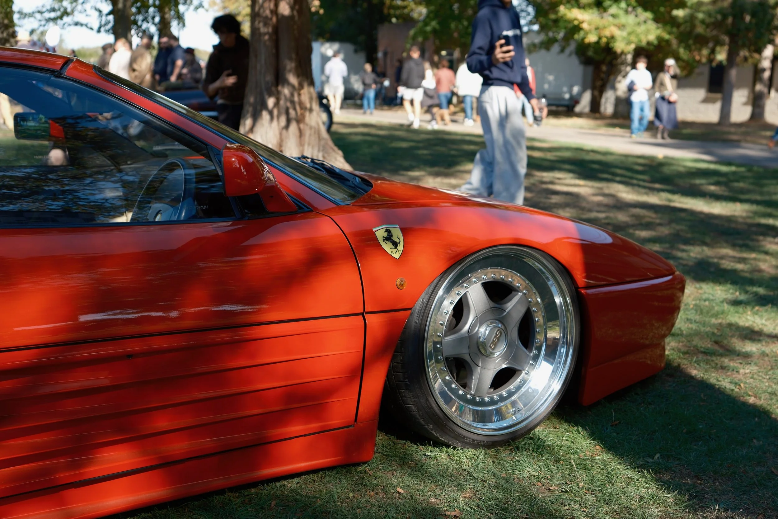 Close-up of a red Ferrari sports car with shiny silver wheels, parked on grass during a car show with people walking and browsing in the background.