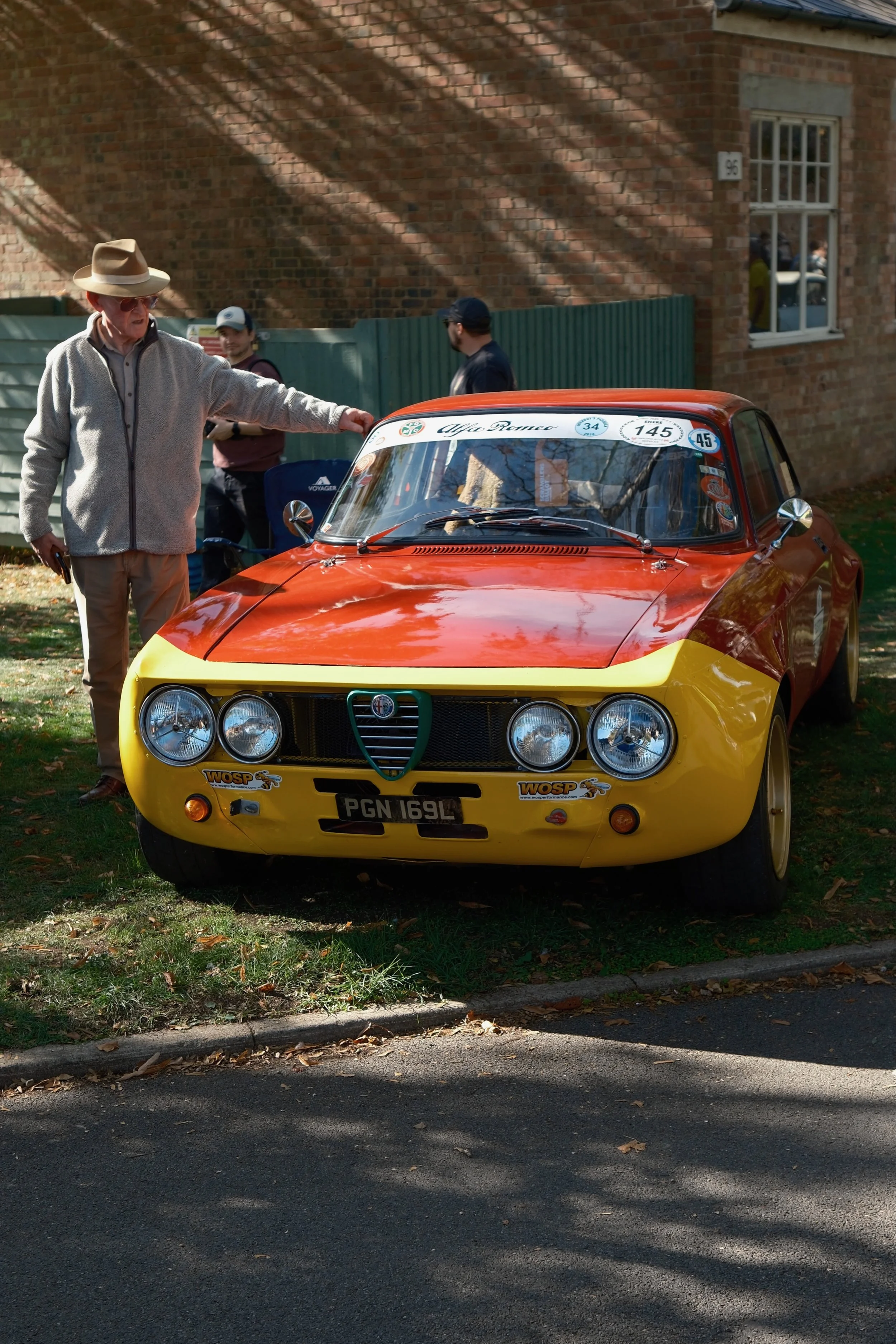 A vintage orange and yellow racing car parked on a grassy area beside a brick building with a window. An older man in a light hat and sweater is touching the car, with three other people in the background.