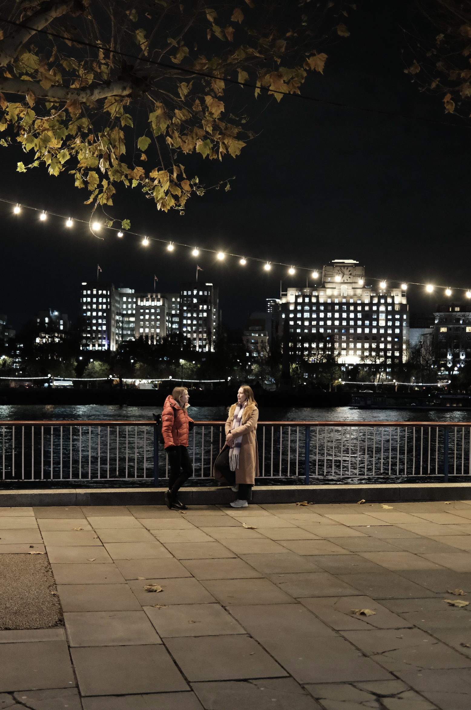 Two women standing on a paved walkway beside a waterway at night, with illuminated city buildings and string lights overhead, and a tree with autumn leaves in the foreground.