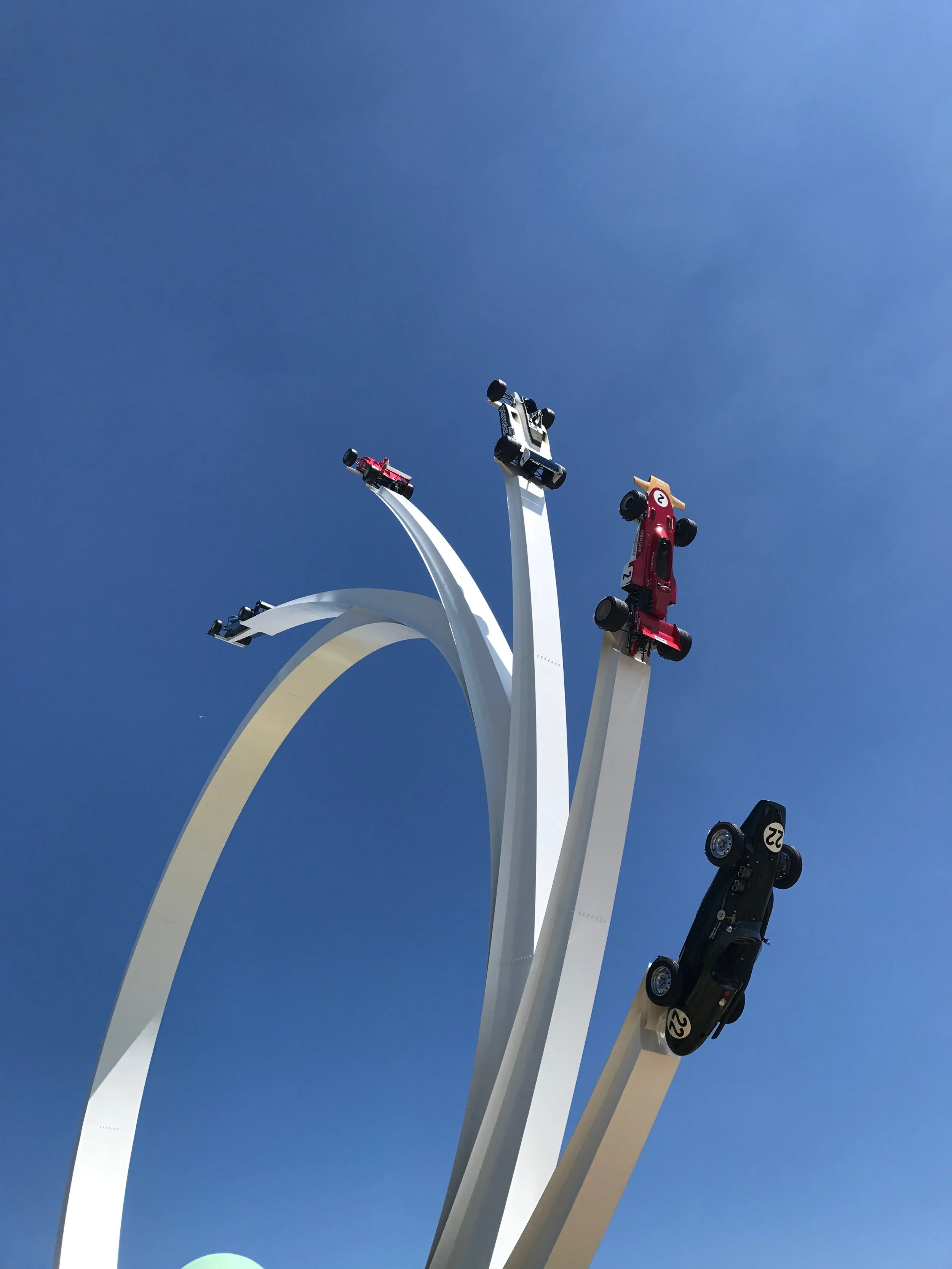 Close-up view of the top of the Cloud Gate sculpture in Chicago, with several toy cars attached to rods that emerge from the top of the sculpture against a clear blue sky.
