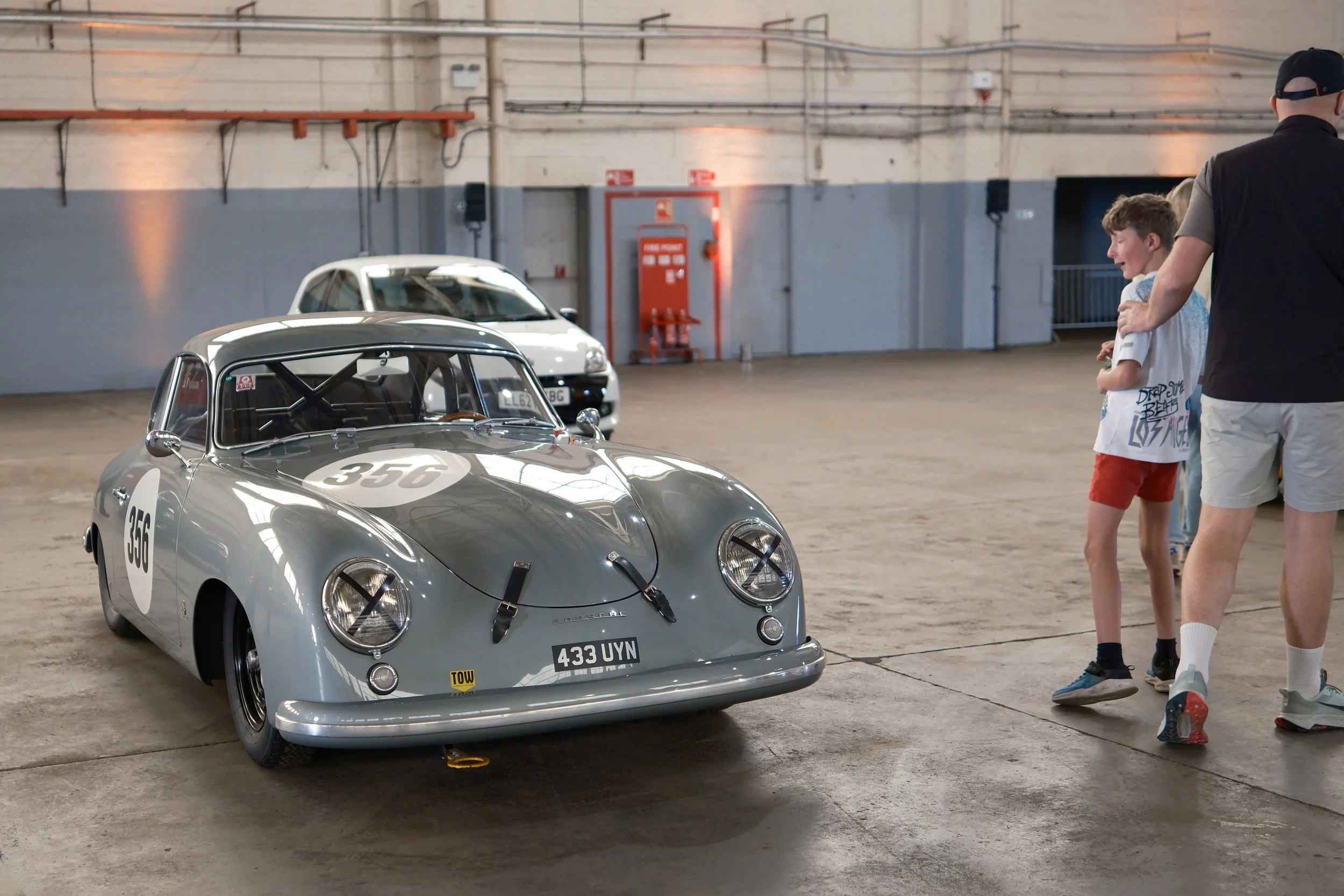 A vintage race car with the number 356 on its hood and sides is parked inside an industrial building, with two young boys and an adult man standing nearby, looking at the car.