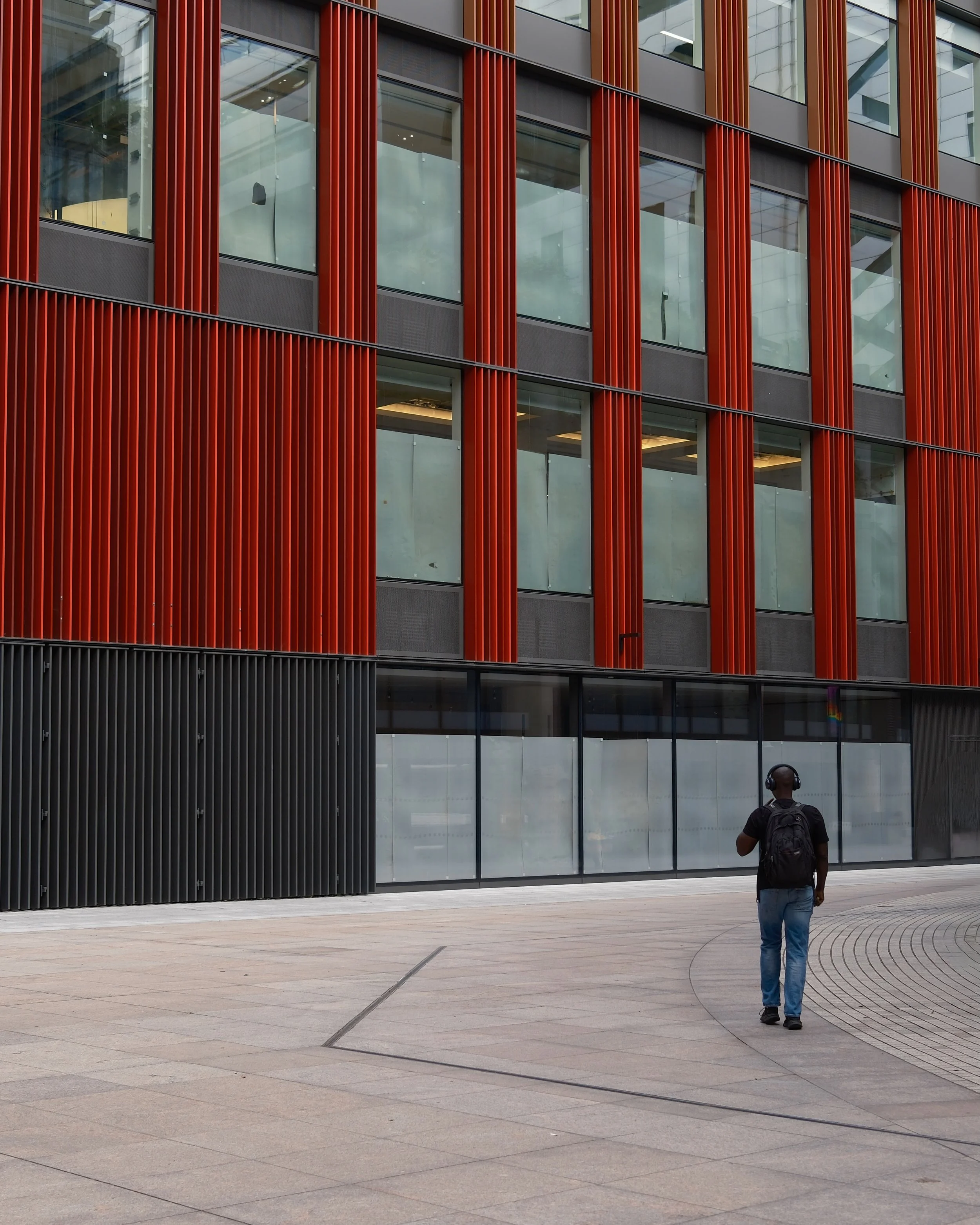 A person with a backpack and headphones walking towards a modern building with red vertical panels and large glass windows.