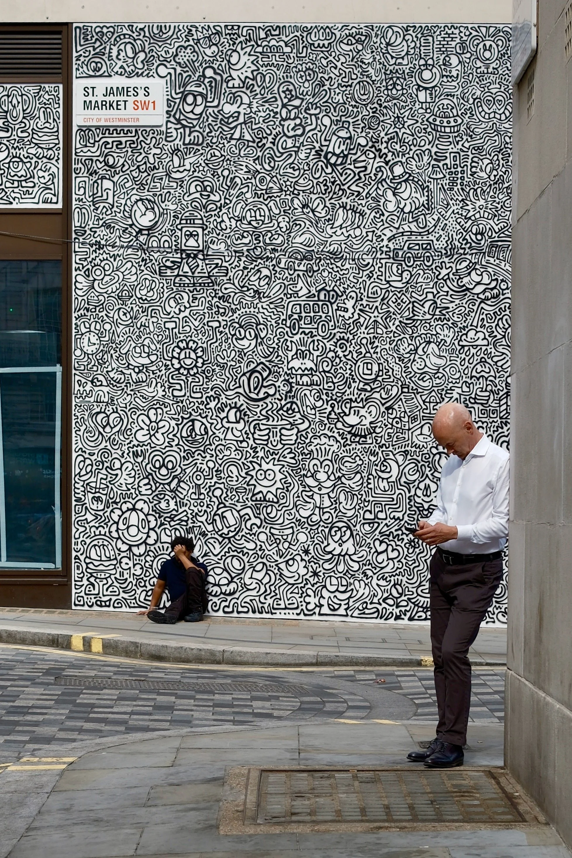 Street scene with a large black-and-white mural filled with cartoonish doodles. Two people are in the scene: a young person sitting on the ground with a backpack and a man standing nearby, looking at a phone. The street has a curb, a grate, and textu