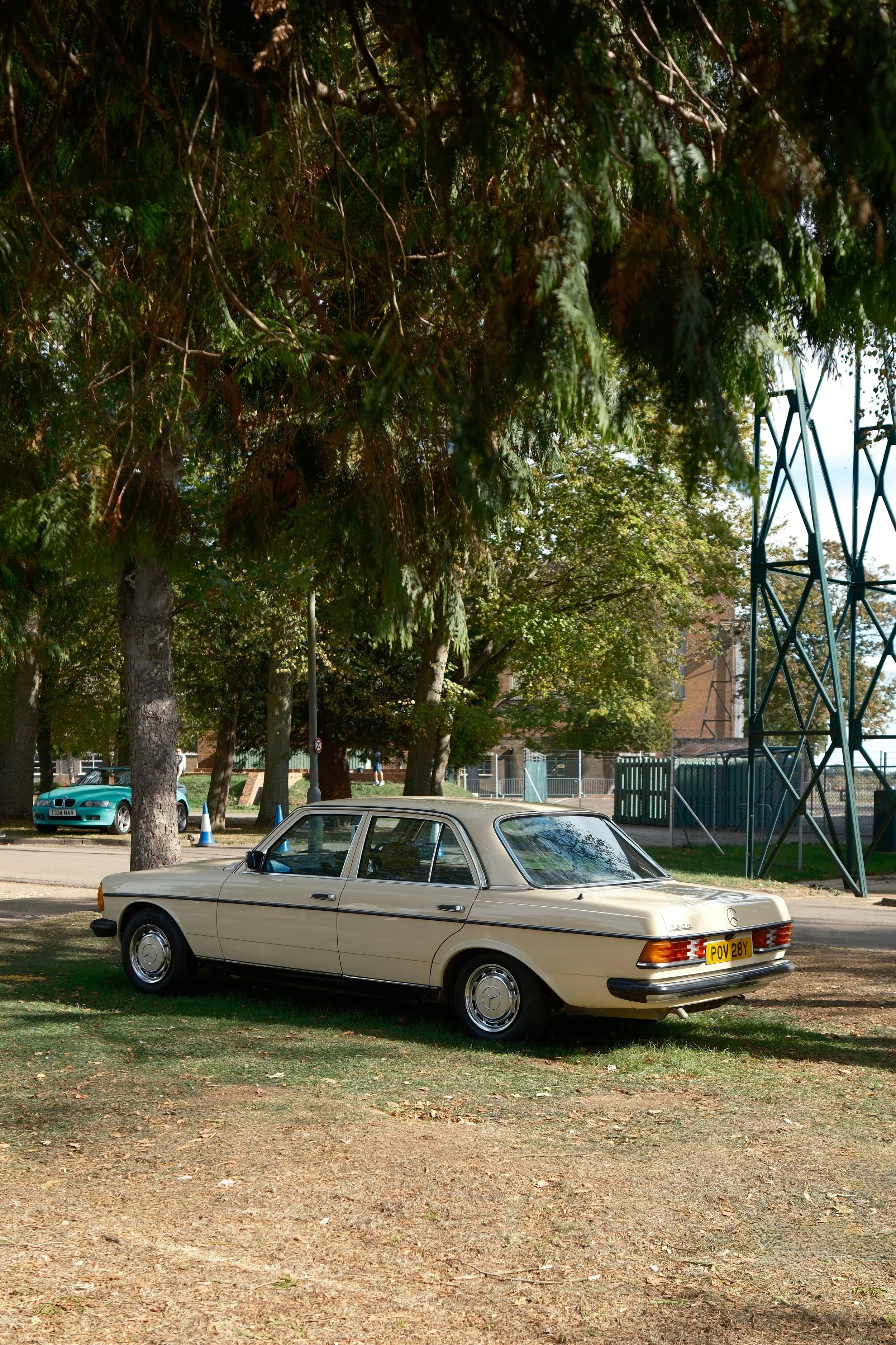 A vintage beige Mercedes-Benz car parked on a grassy area with trees and a metal fence in the background.